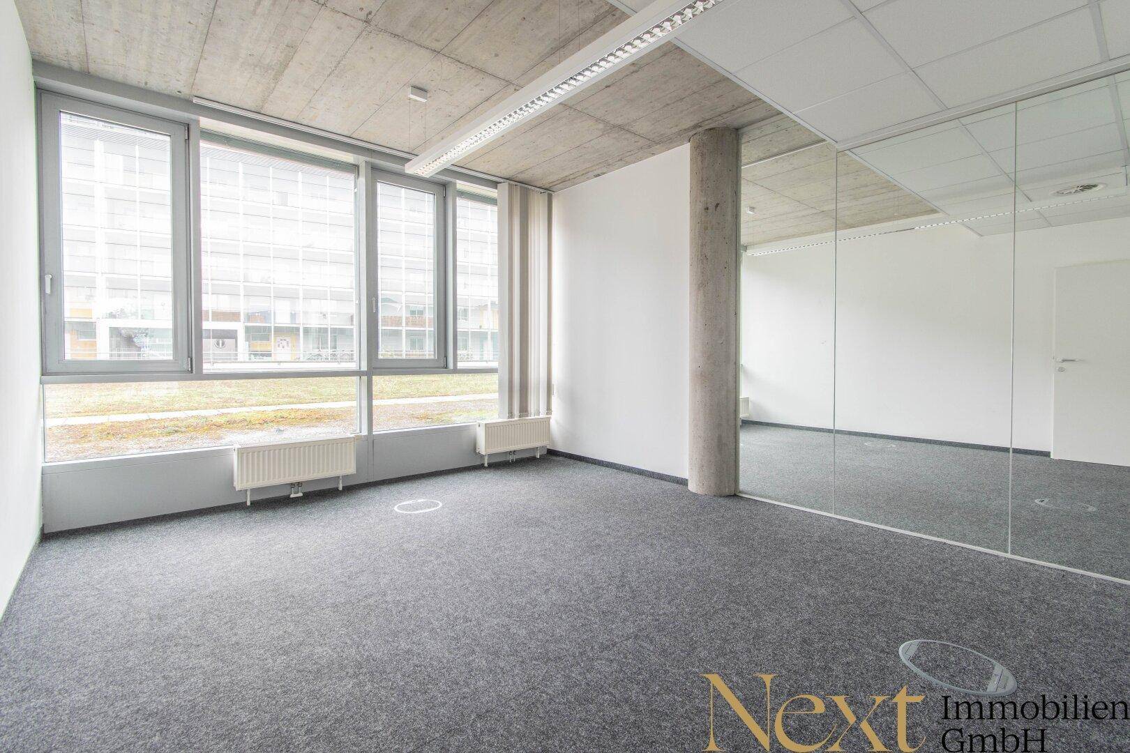 Helle Büroecke mit großen Fenstern, grauem Teppichboden, Sichtbetondecke und Spiegelfläche.
