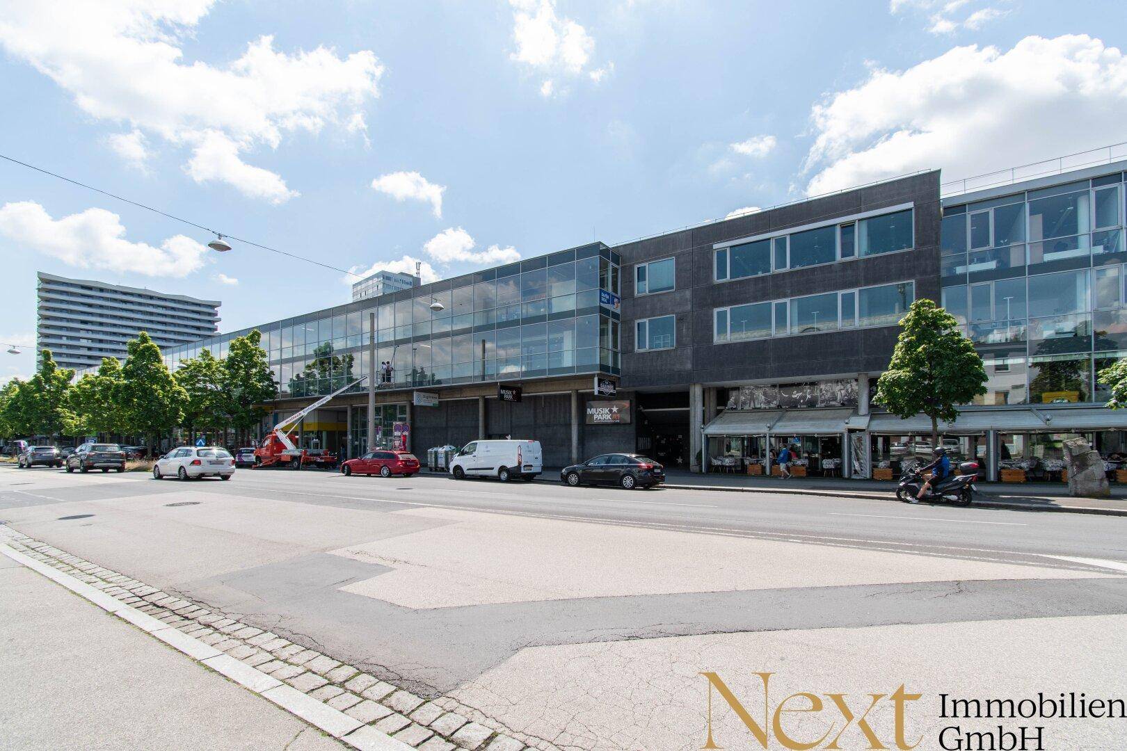 Langes Geschäftsgebäude entlang einer belebten Straße mit vielen Parkplätzen und Geschäften im Erdgeschoss.