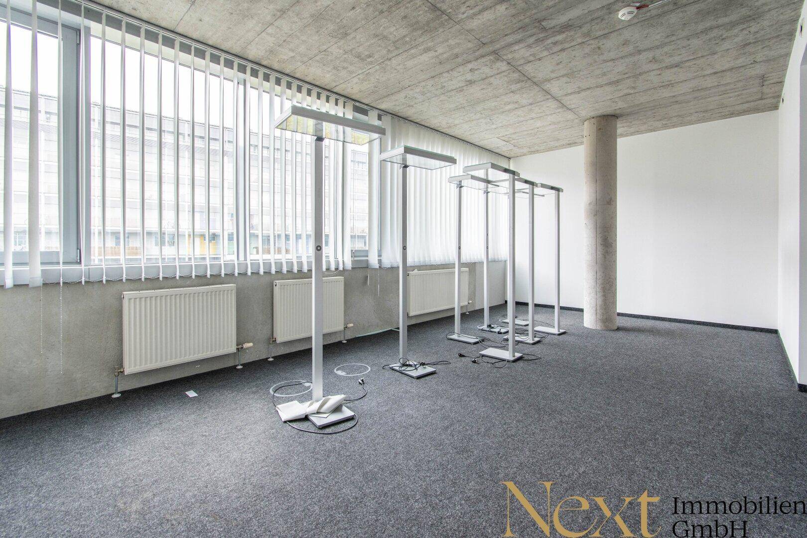 Heller Büroraum mit großen Fenstern, Jalousien und sichtbarer Betondecke, ideal für kreative Arbeitsbereiche.