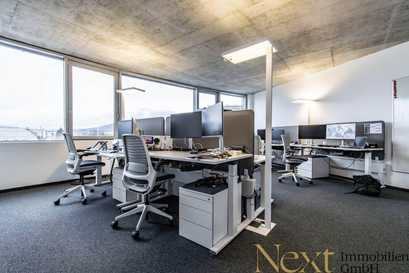 Ein heller, moderner Büroraum mit mehreren Arbeitsplätzen, großen Fenstern und Teppichboden.
