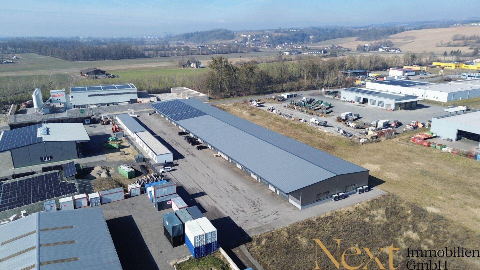 Übersicht aus der Vogelperspektive auf ein Gewerbegebiet mit Hallen, Freiflächen und umliegender grüner Landschaft.