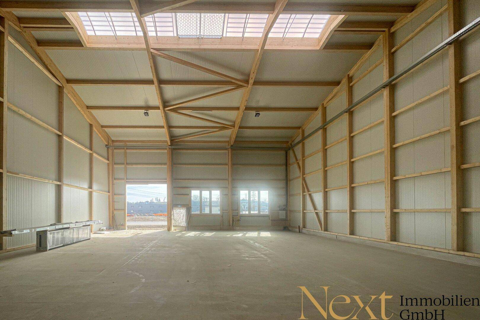 Weitläufige, helle Lagerhalle mit hohen Decken, Holzbalken und großem Tor sowie Fenstern am Ende.
