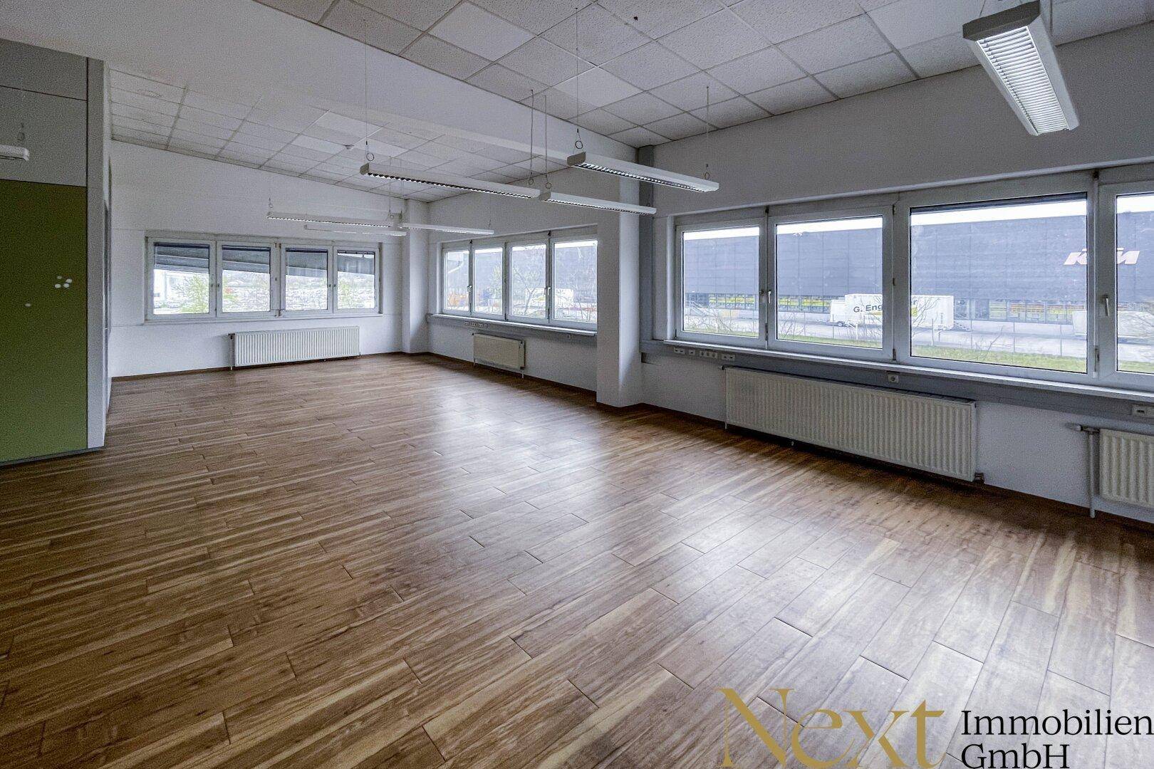 Helles Büro mit großen Fenstern, Holzfußboden und Blick auf das Außengelände.