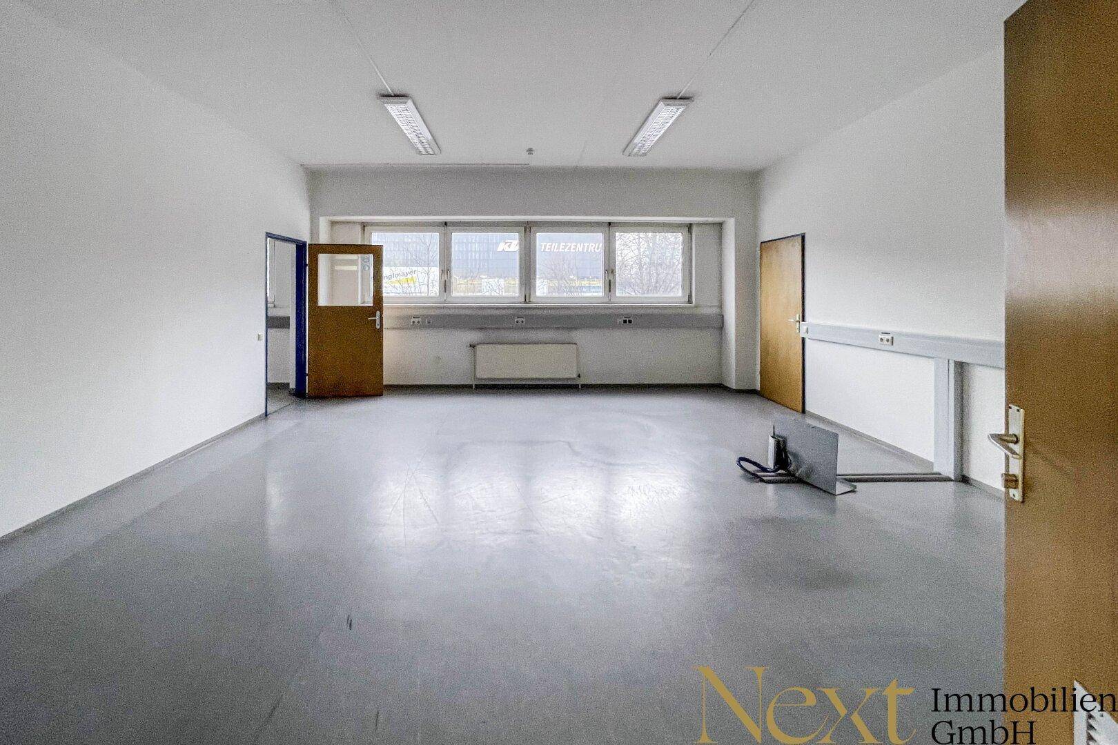 Geräumiges Büro mit grauem Vinylboden und großen Fenstern, ideal für Arbeitsbereiche.