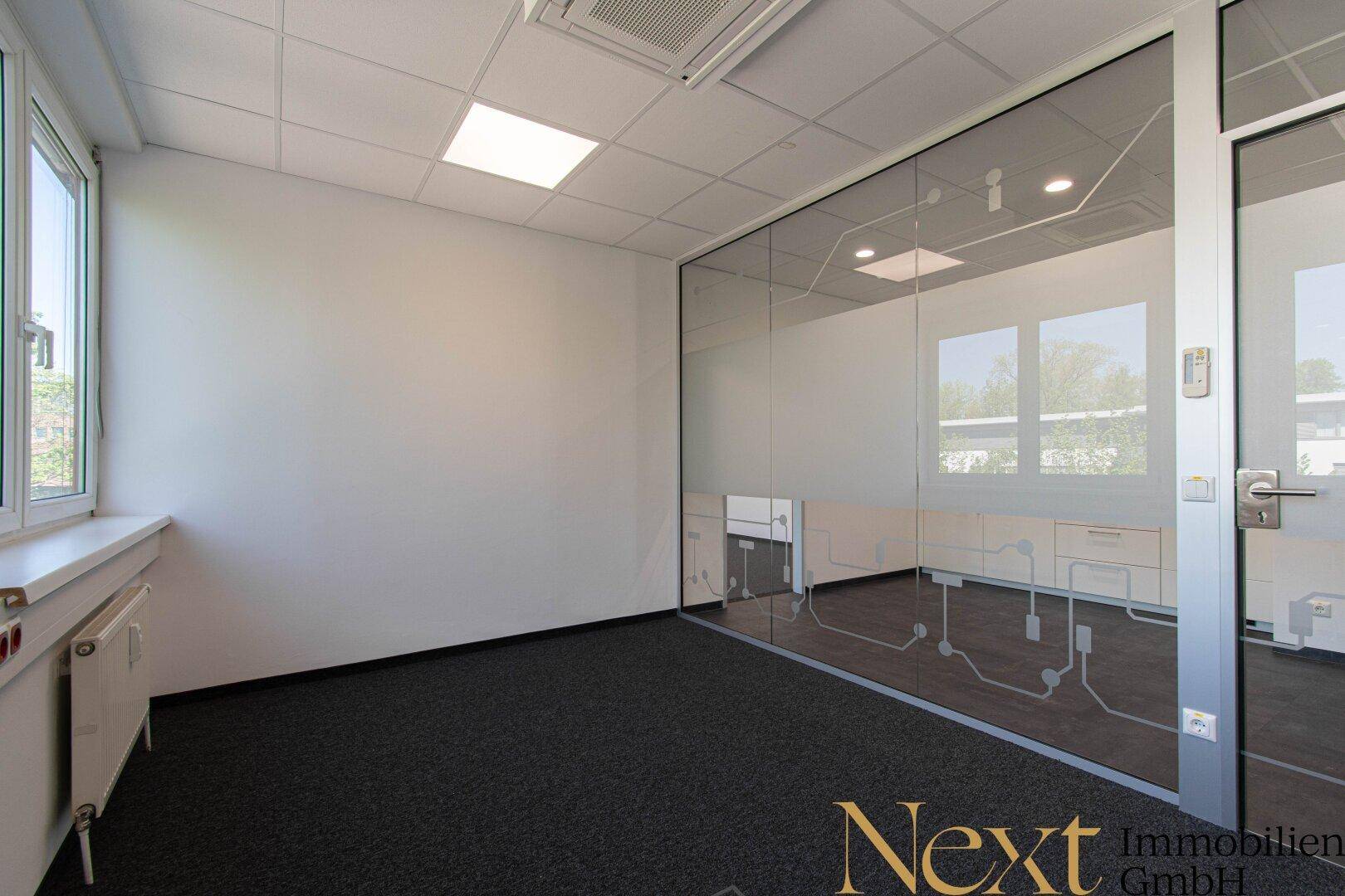 Modernes Büro mit Glaswand, Teppichboden und großen Fenstern, die eine helle und offene Atmosphäre schaffen.