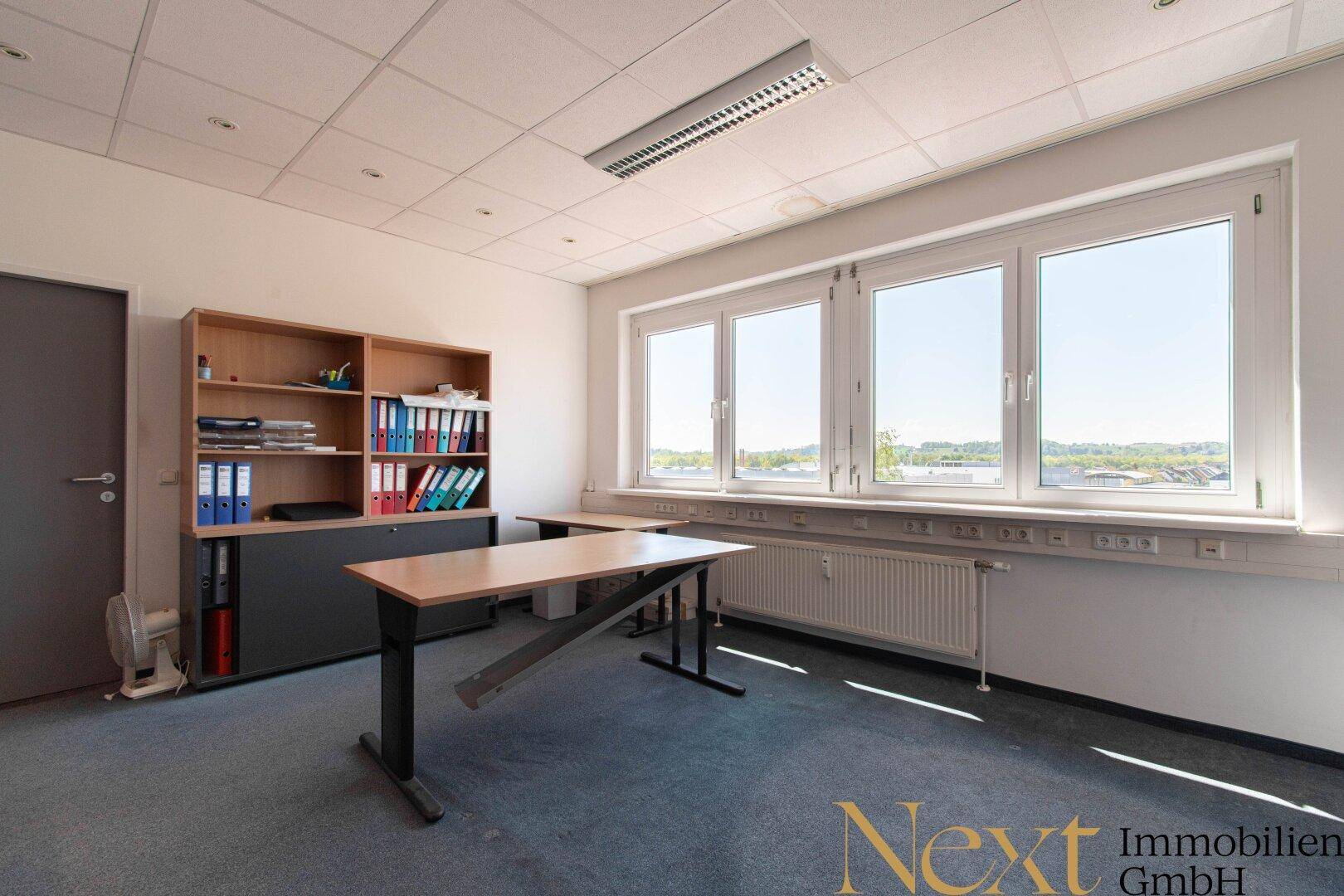 Leeres Büro mit Teppichboden, großen Fenstern und Heizkörper unter der Fensterbank, bereit zur Nutzung.