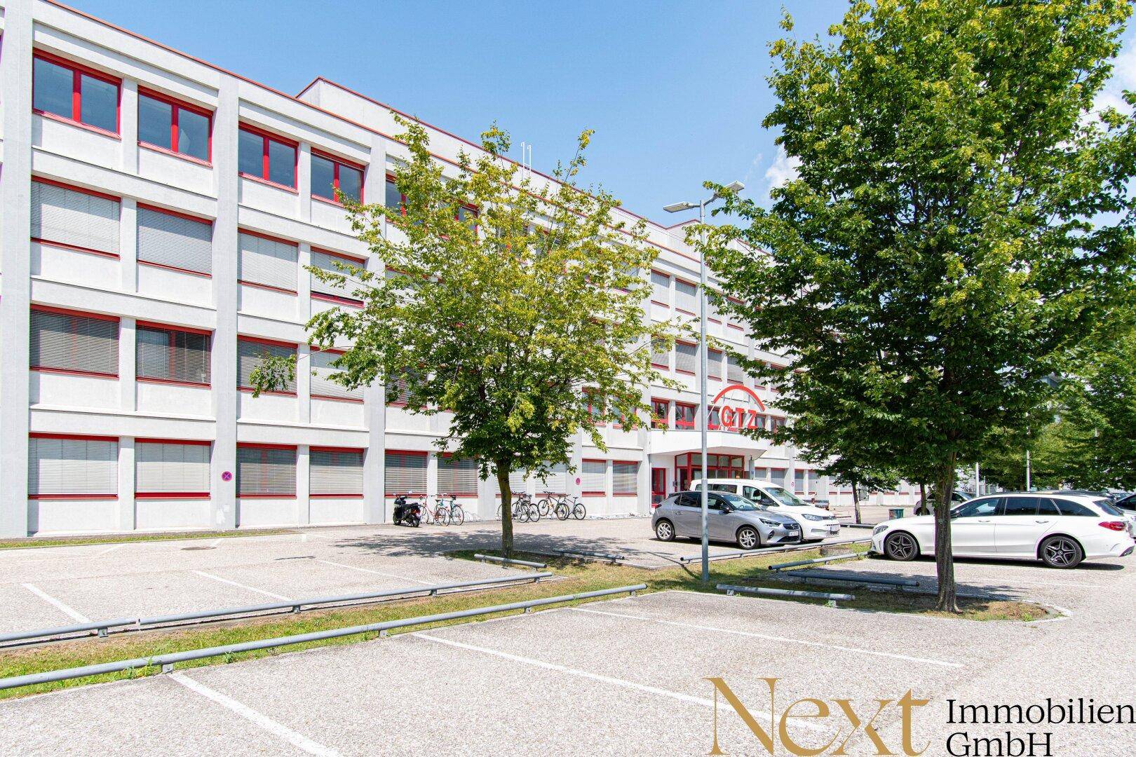 Ansicht des Bürogebäudes von außen mit Parkplätzen und Bäumen im Vordergrund an einem sonnigen Tag.