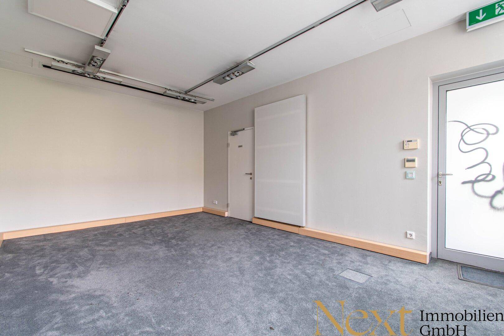 Leerer Gewerberaum mit grauem Teppichboden, hellen Wänden und sichtbaren Deckeninstallationen.