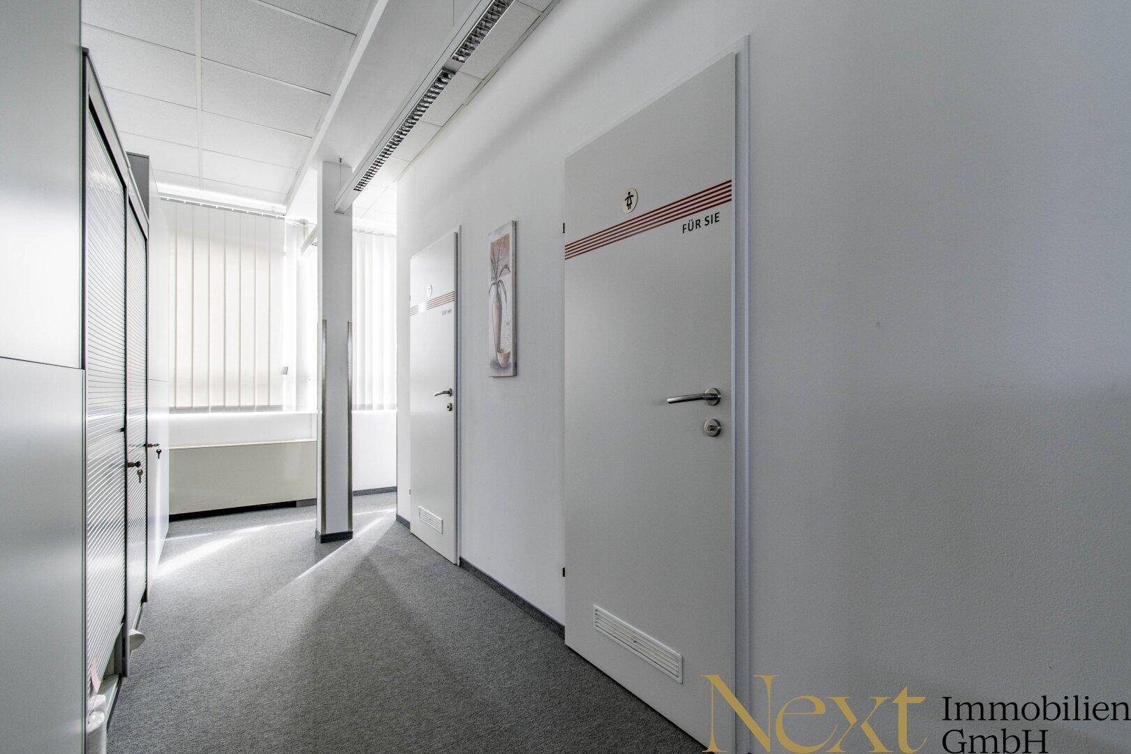 Langer, heller Flur in einem Bürogebäude mit mehreren Türen und einem Fenster mit Jalousien.