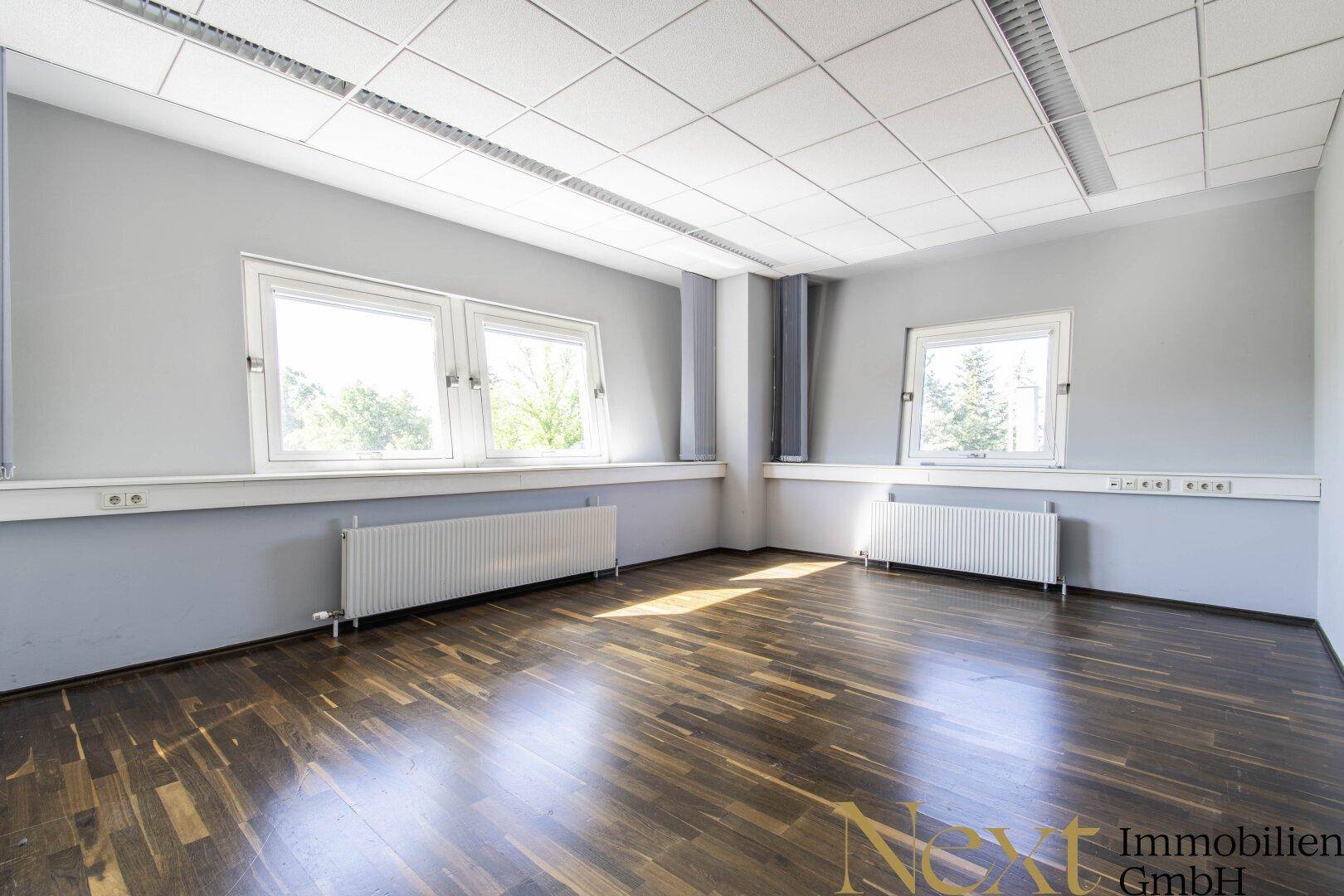 Helles Büro mit dunklem Holzboden, grauen Wänden und großen Fenstern, die eine angenehme Arbeitsatmosphäre schaffen.