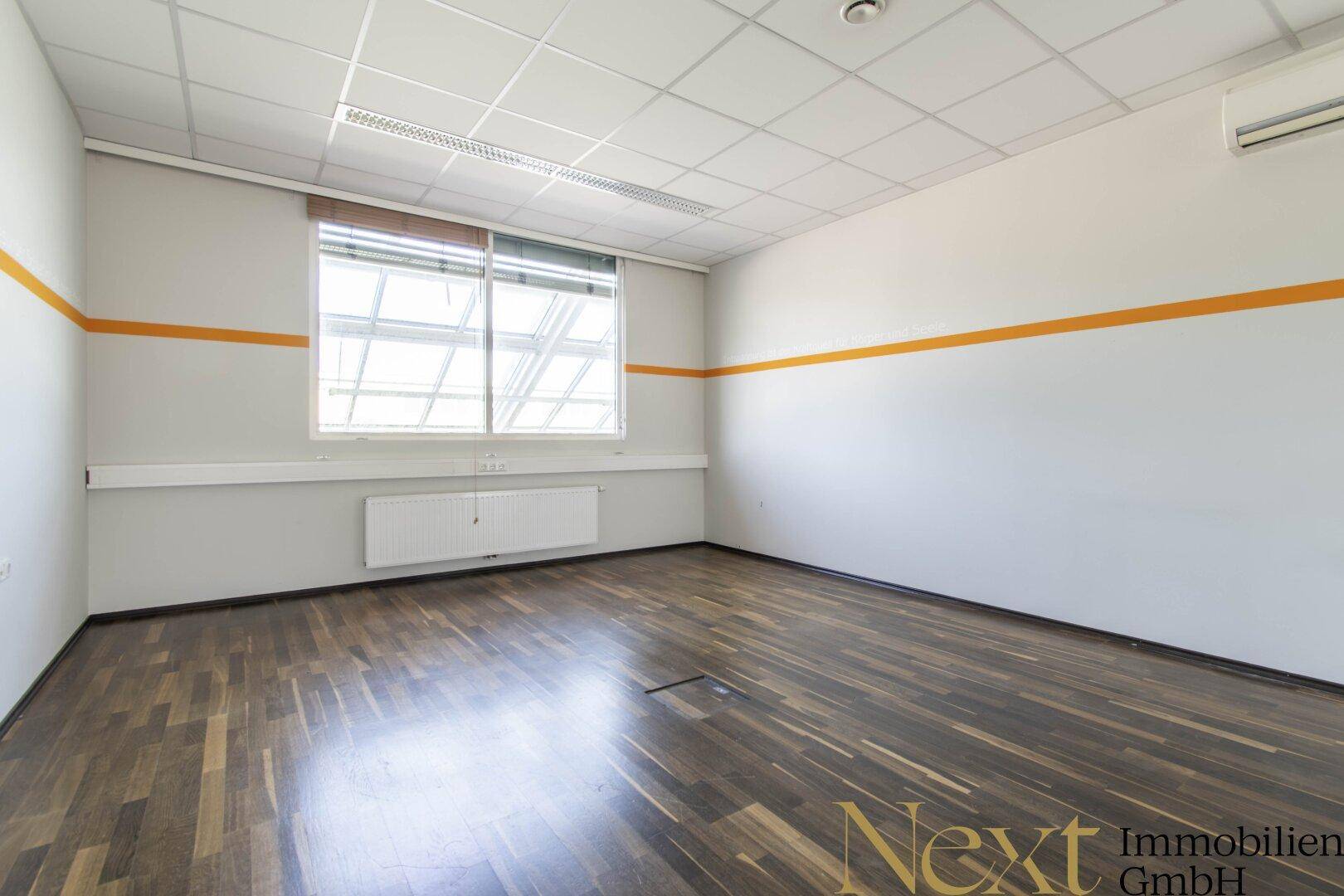 Büro mit Holzboden, weißen Wänden und einem orangefarbenen Streifen, ausgestattet mit Klimaanlage und Fenster.