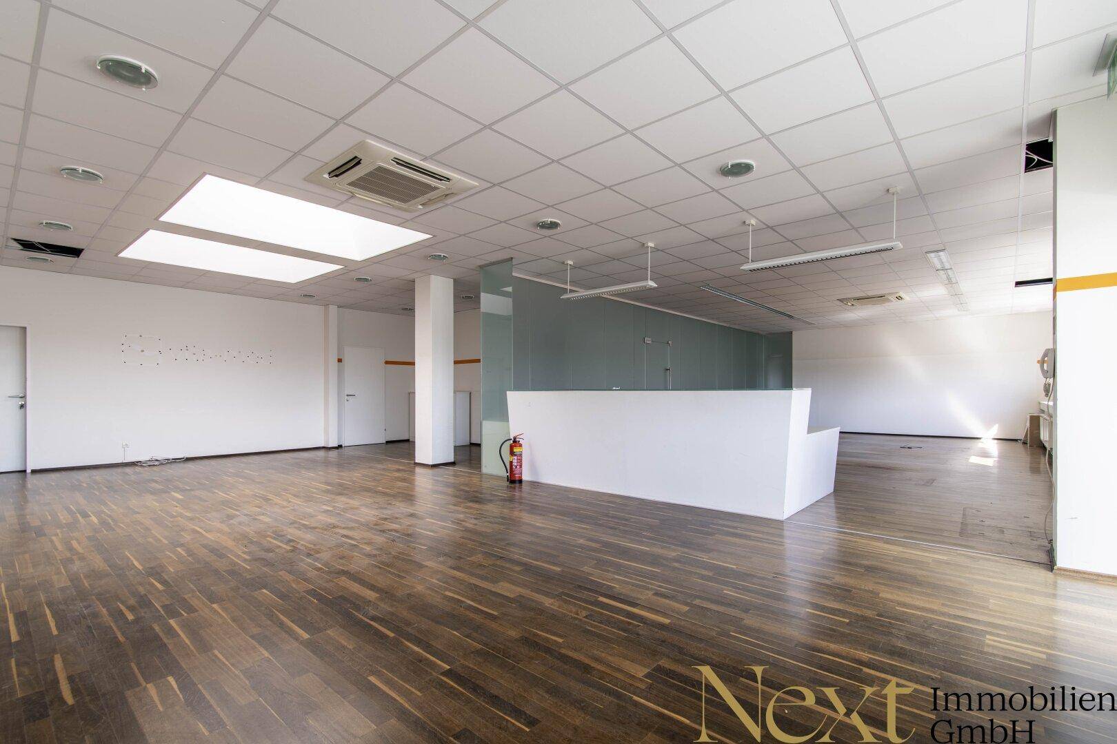 Großer offener Raum mit Holzboden, Empfangsbereich und Oberlichtern, ideal für Einzelhandel oder Showroom.