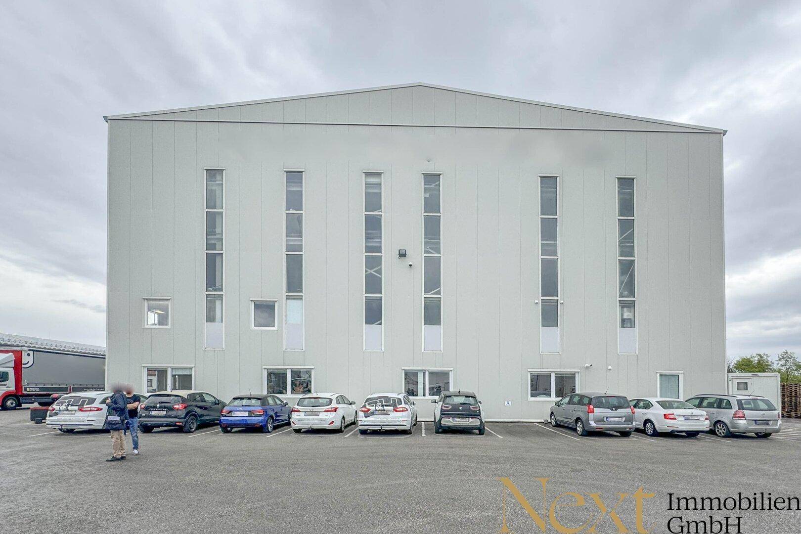 Moderne Gewerbeimmobilie mit großer Fassade, hohen Fenstern und Parkplätzen davor.