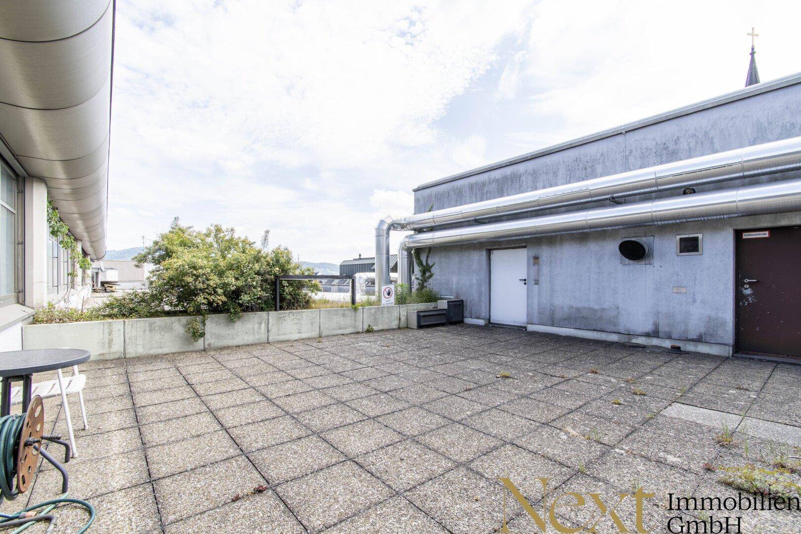 Große Dachterrasse mit Betonplatten, etwas Grün und Blick auf die Umgebung, ideal für Pausen.