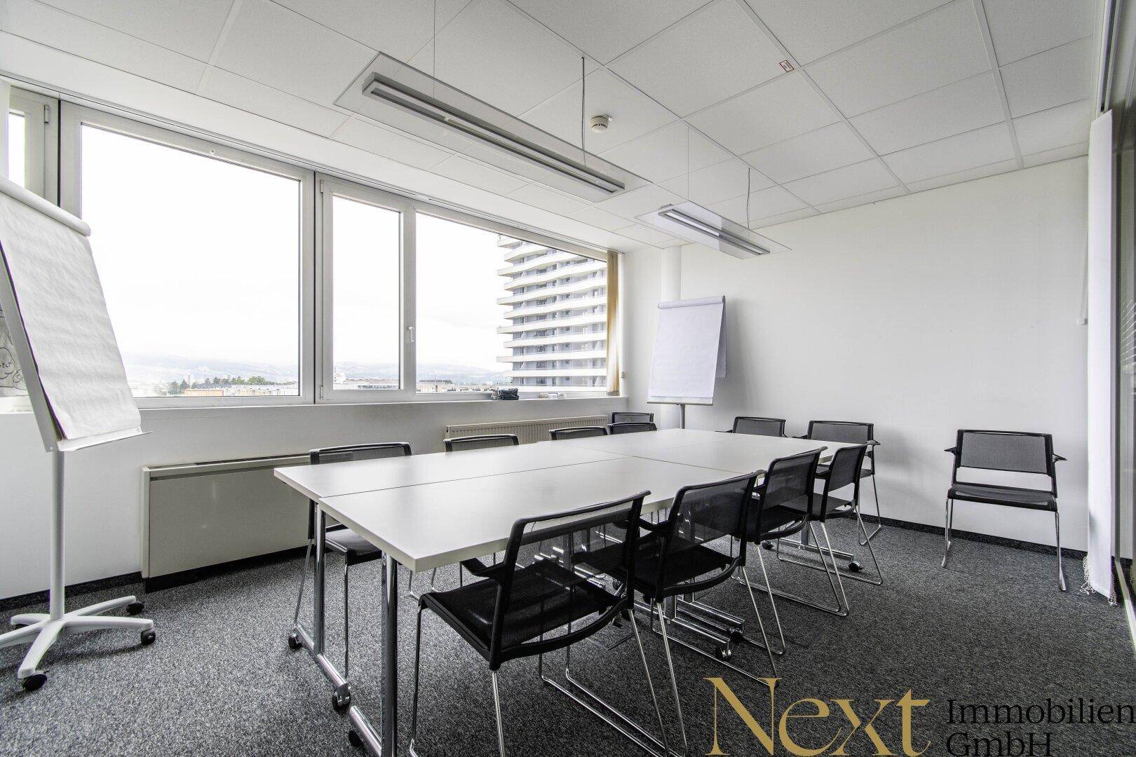 Heller Konferenzraum mit großem Tisch, Flipcharts und Panoramablick durch die Fensterfront.