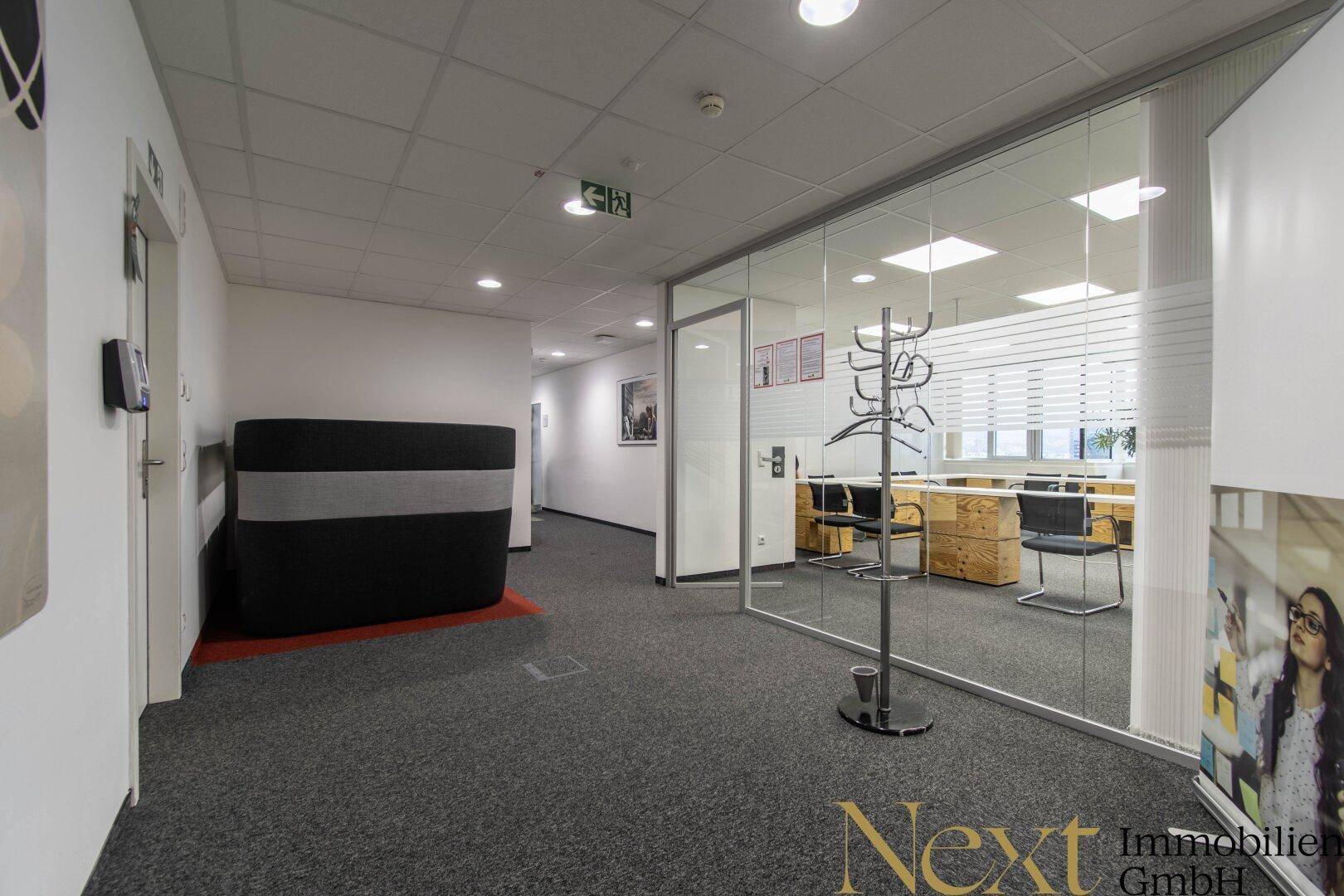 Heller Bürogang mit Glaswand, die Einblick in einen modernen Bürobereich gewährt, und einer Sitzgelegenheit.