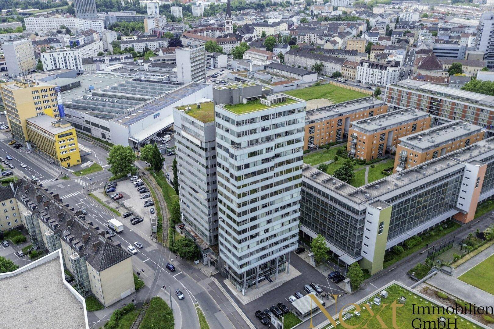 Luftaufnahme eines modernen Gebäudekomplexes in einer städtischen Umgebung mit viel Grün und umliegenden Straßen.
