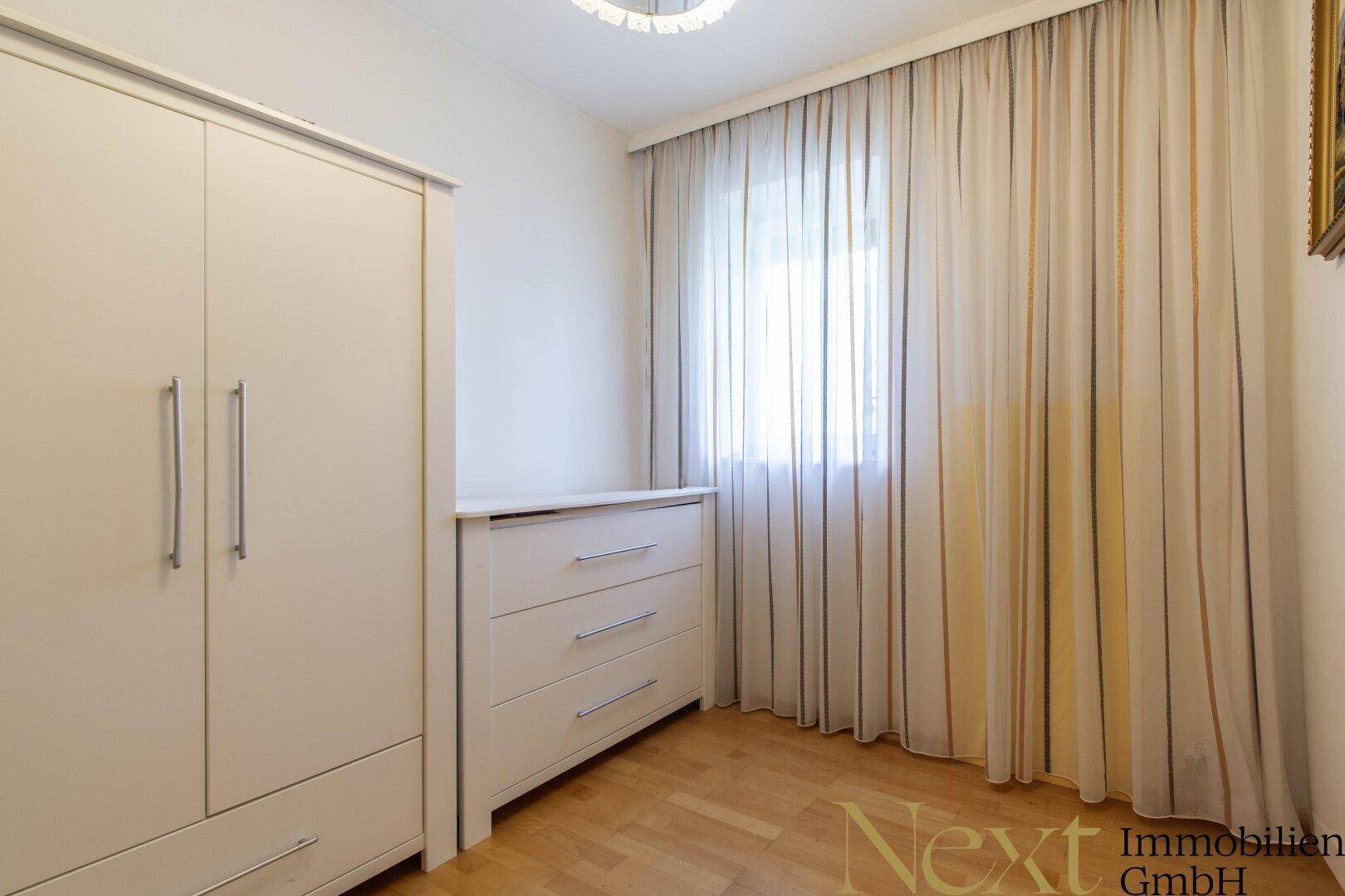 Helles Schlafzimmer mit Kleiderschrank, Kommode und Fenster mit Gardinen.