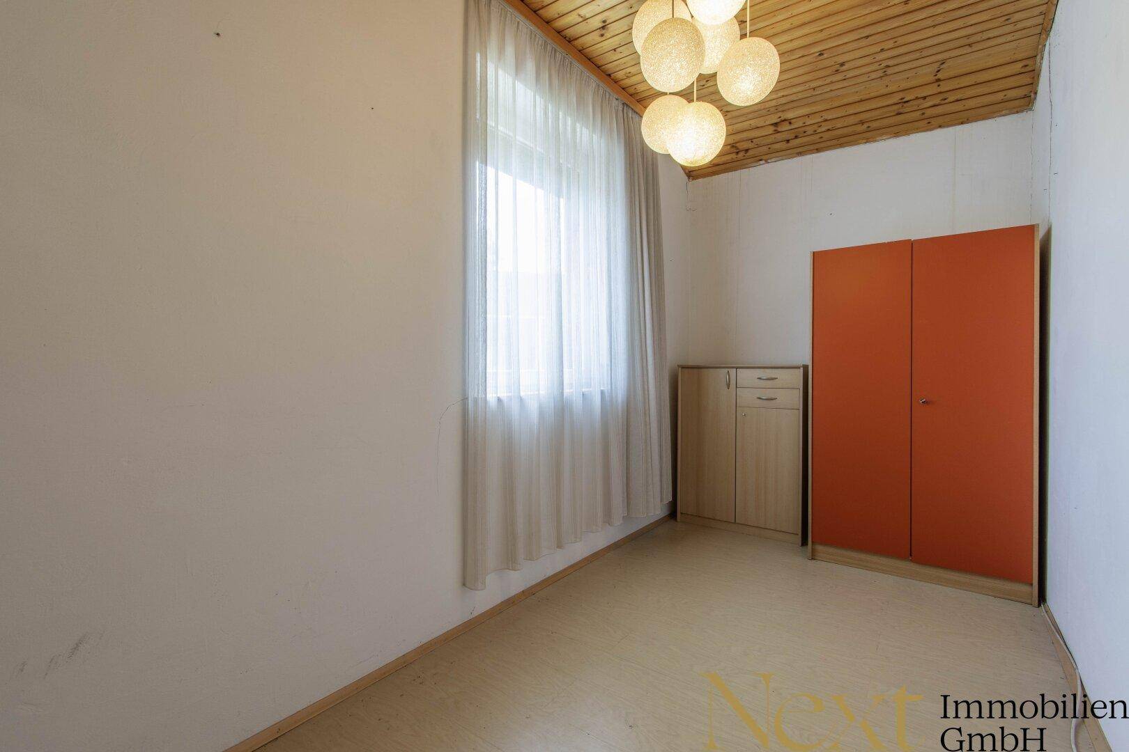 Einfaches Schlafzimmer mit Holzdecke, Fenster und orangefarbenem Schrank.
