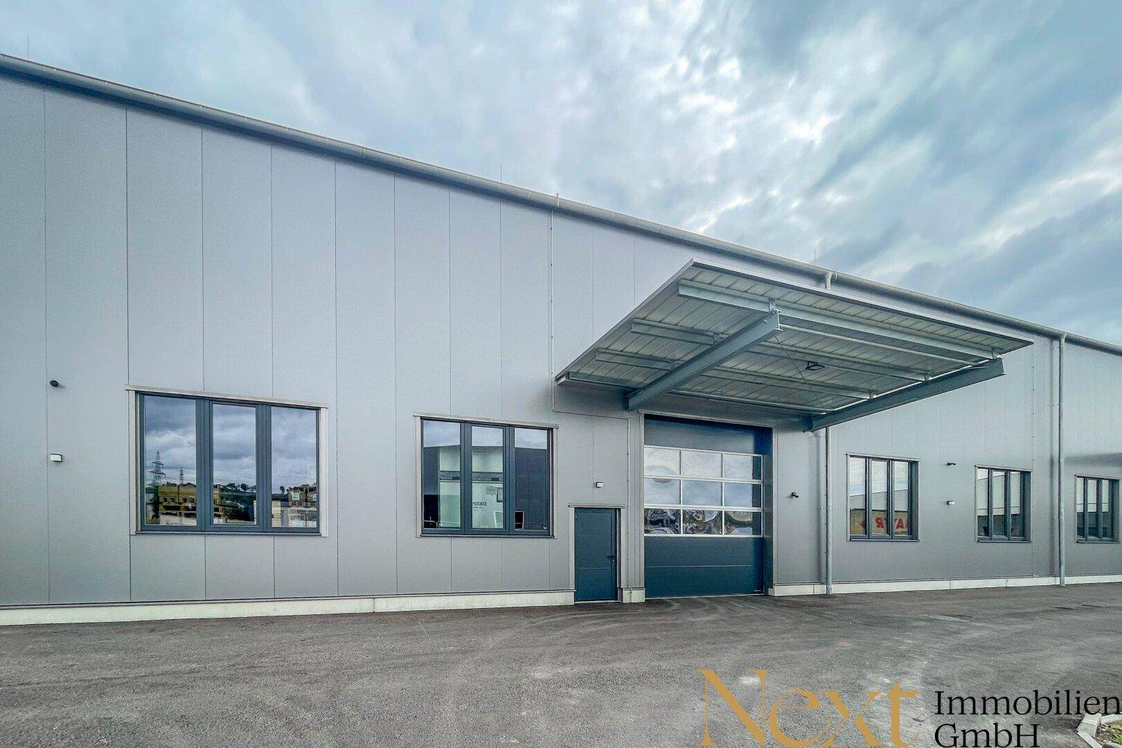 Moderne Industriefassade mit grauen Metallpaneelen, großen Fenstern und einem überdachten Rolltor.