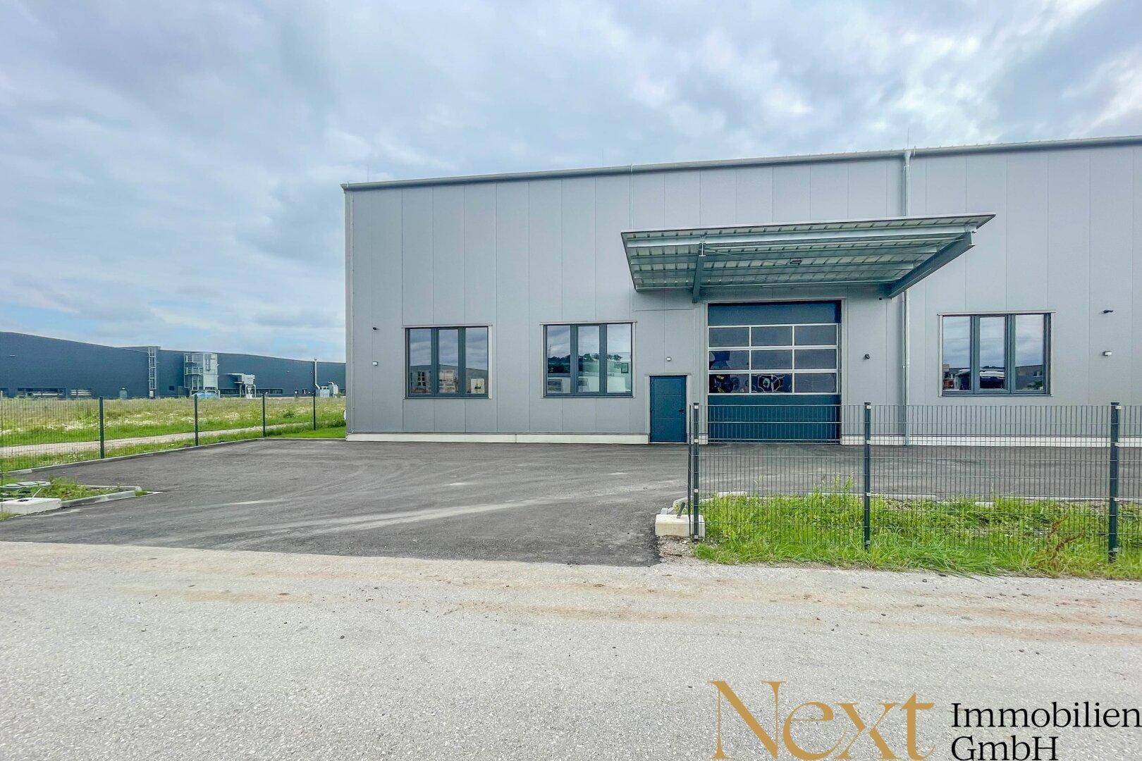 Außenansicht des modernen Industriegebäudes mit Zufahrt, Zaun und Blick auf weitere Gewerbebauten.