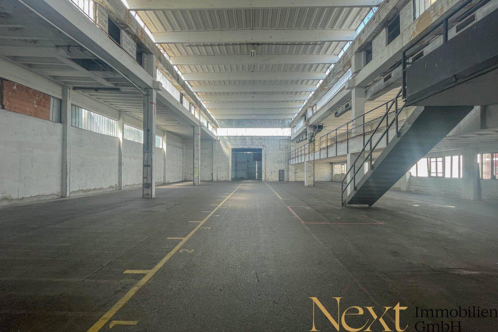 Weitläufige Industriehalle mit hohen Decken, großen Fenstern und einer Treppe zu einer oberen Ebene.