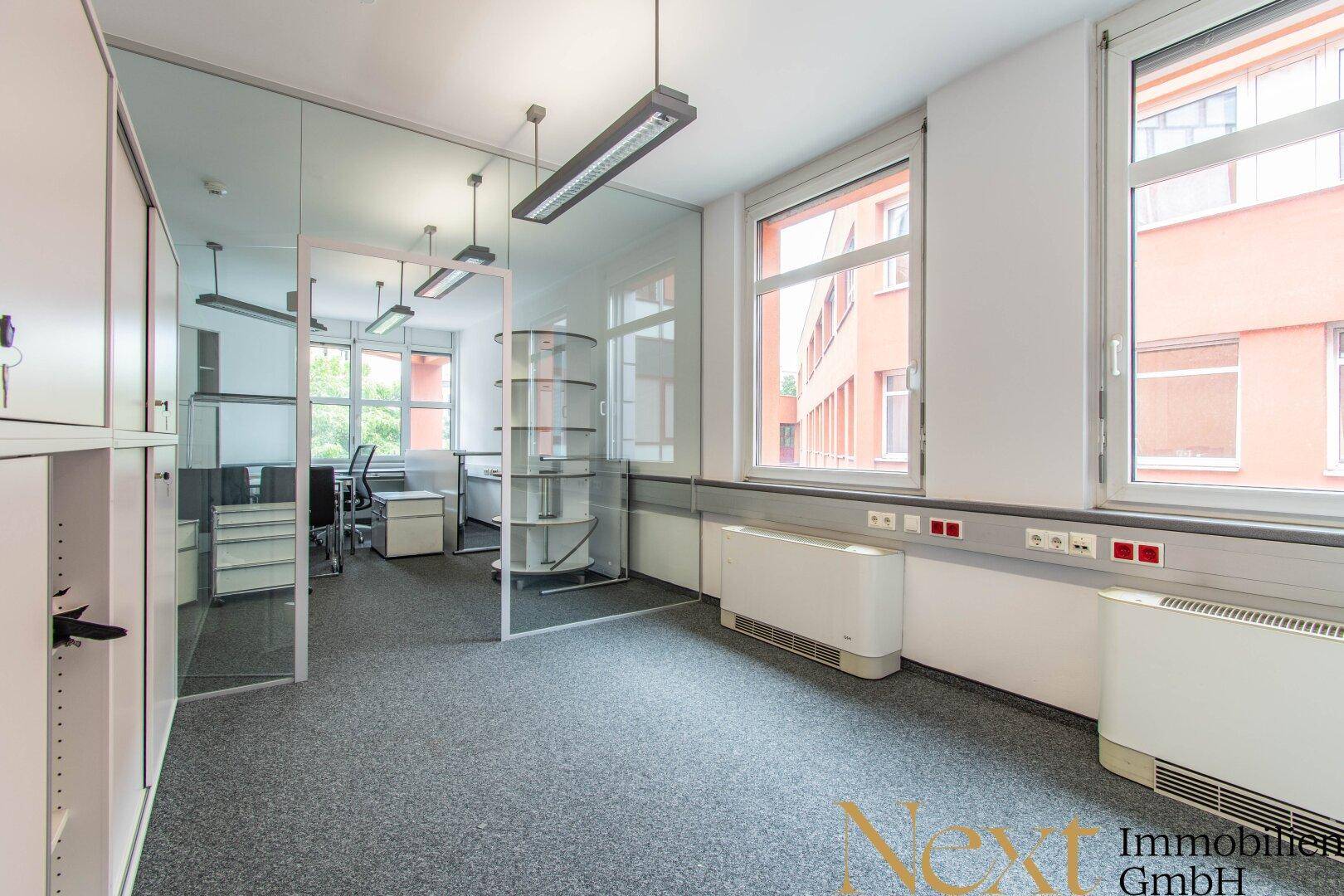 Helles Büro mit Teppichboden, großen Fenstern und einer modernen Glasabtrennung zu einem weiteren Arbeitsbereich.