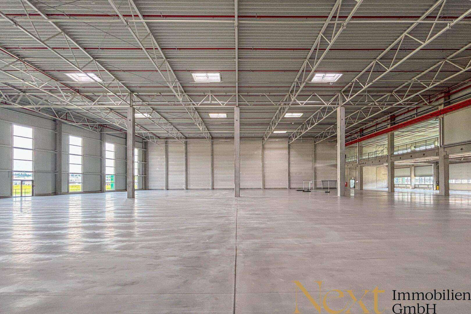 Große, leere Lagerhalle mit hohen Decken, Betonboden und viel Tageslicht durch Fenster und Oberlichter.