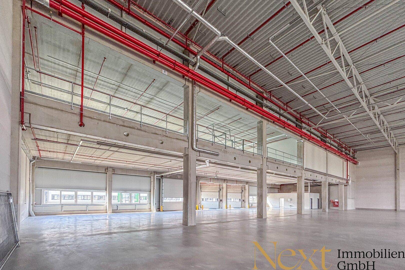 Innenansicht einer modernen Lagerhalle mit Rolltoren, einer oberen Ebene und sichtbaren roten Rohren.