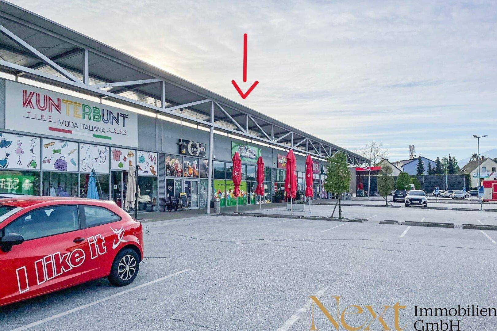 Außenansicht eines modernen Geschäftsgebäudes mit zahlreichen Geschäften und einem großen Parkplatz davor.