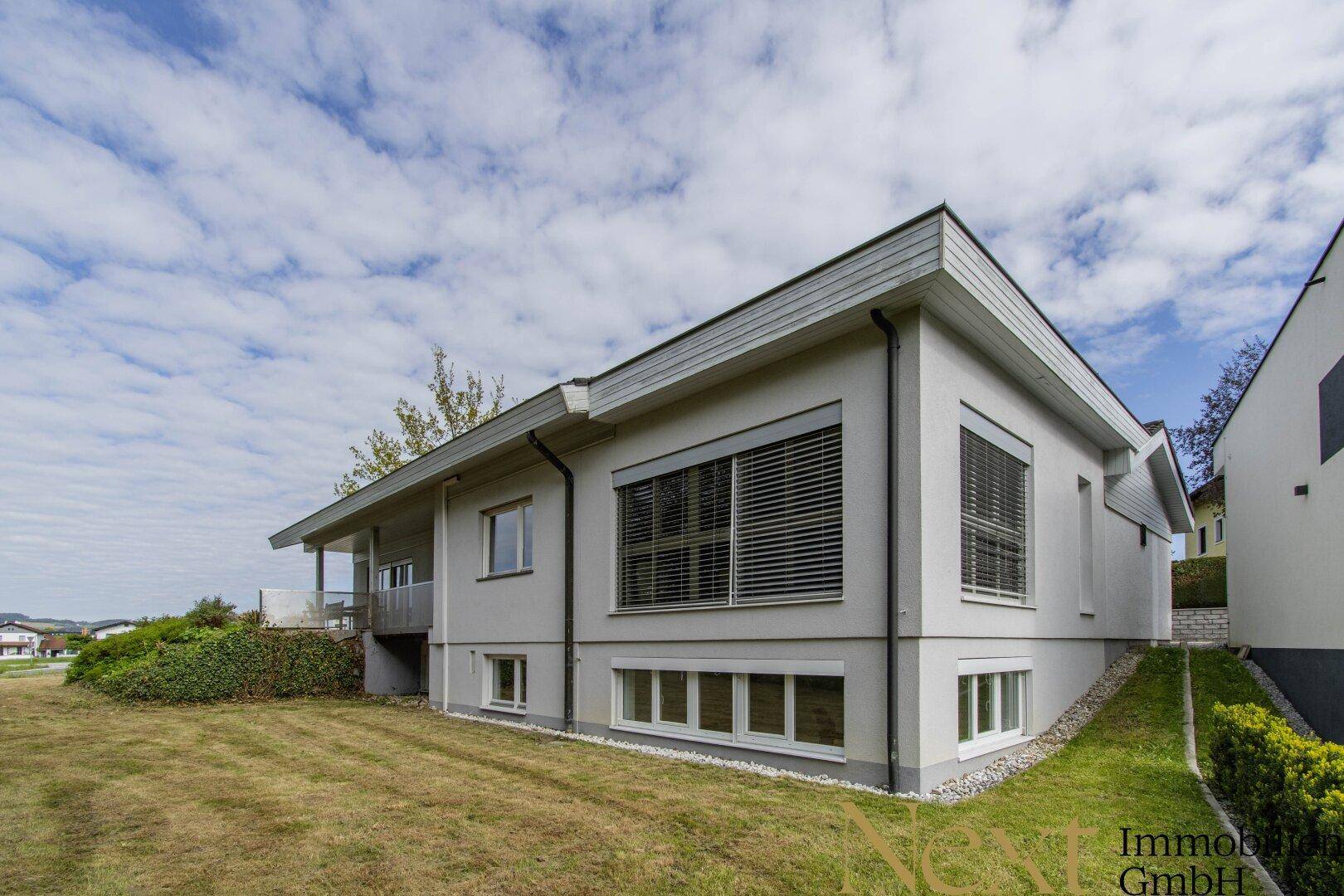 Seitenansicht des modernen Hauses mit großen Fenstern und Jalousien, umgeben von Grünflächen.