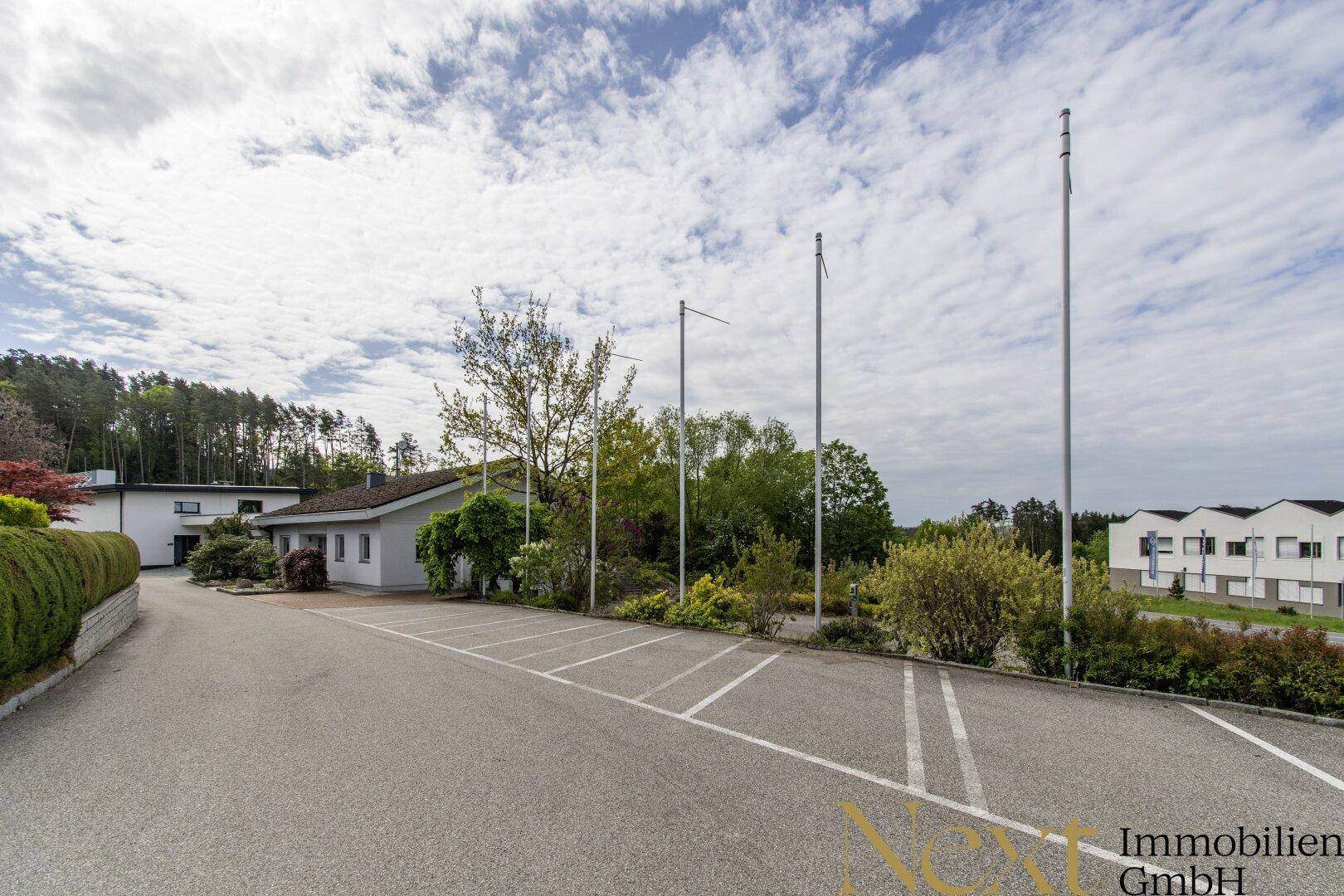 Großer Parkplatz mit markierten Stellflächen und Fahnenmasten vor dem Gebäude in ländlicher Umgebung.