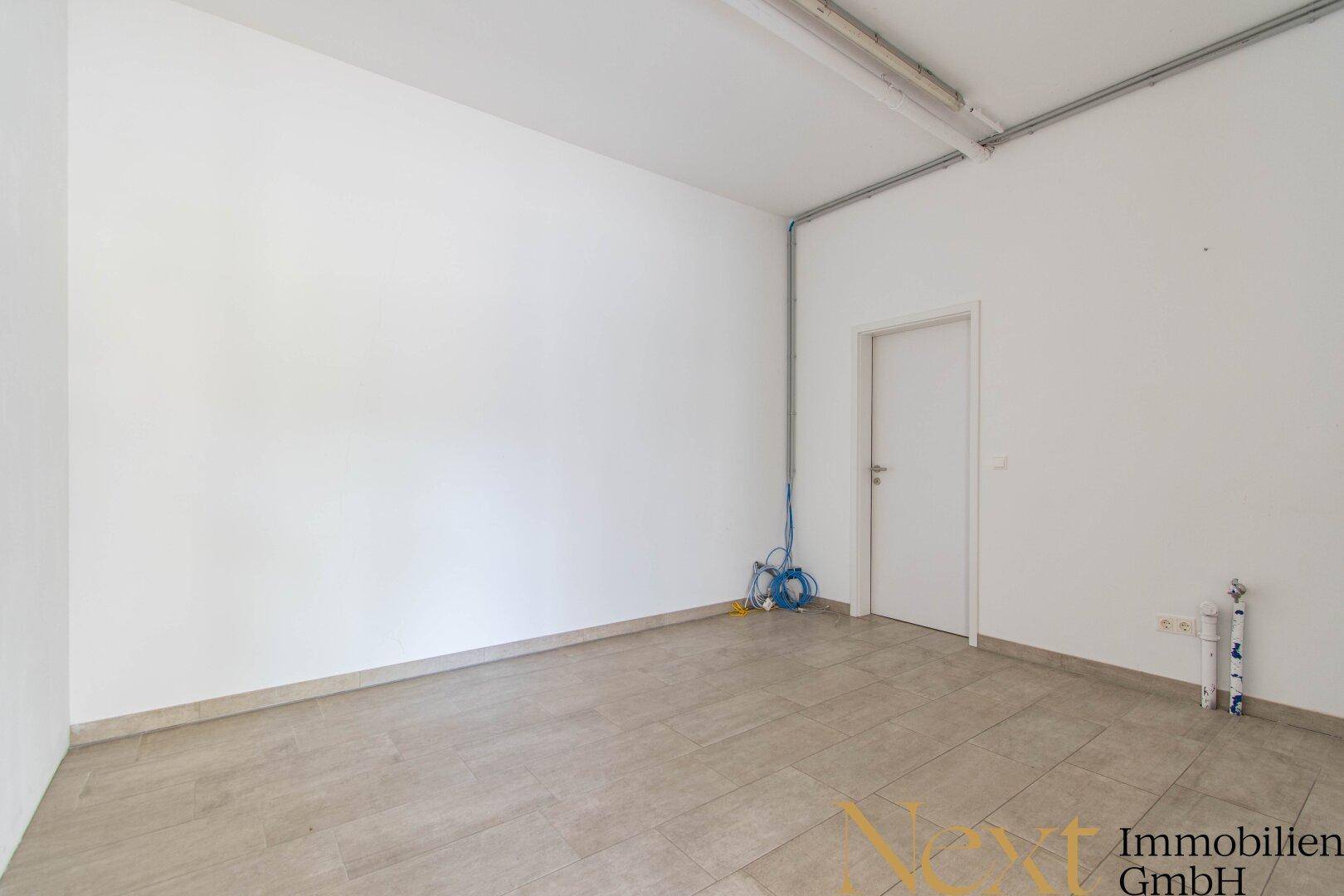 Heller, leerer Raum mit Fliesenboden, einer Tür und sichtbaren Installationen an der Wand.