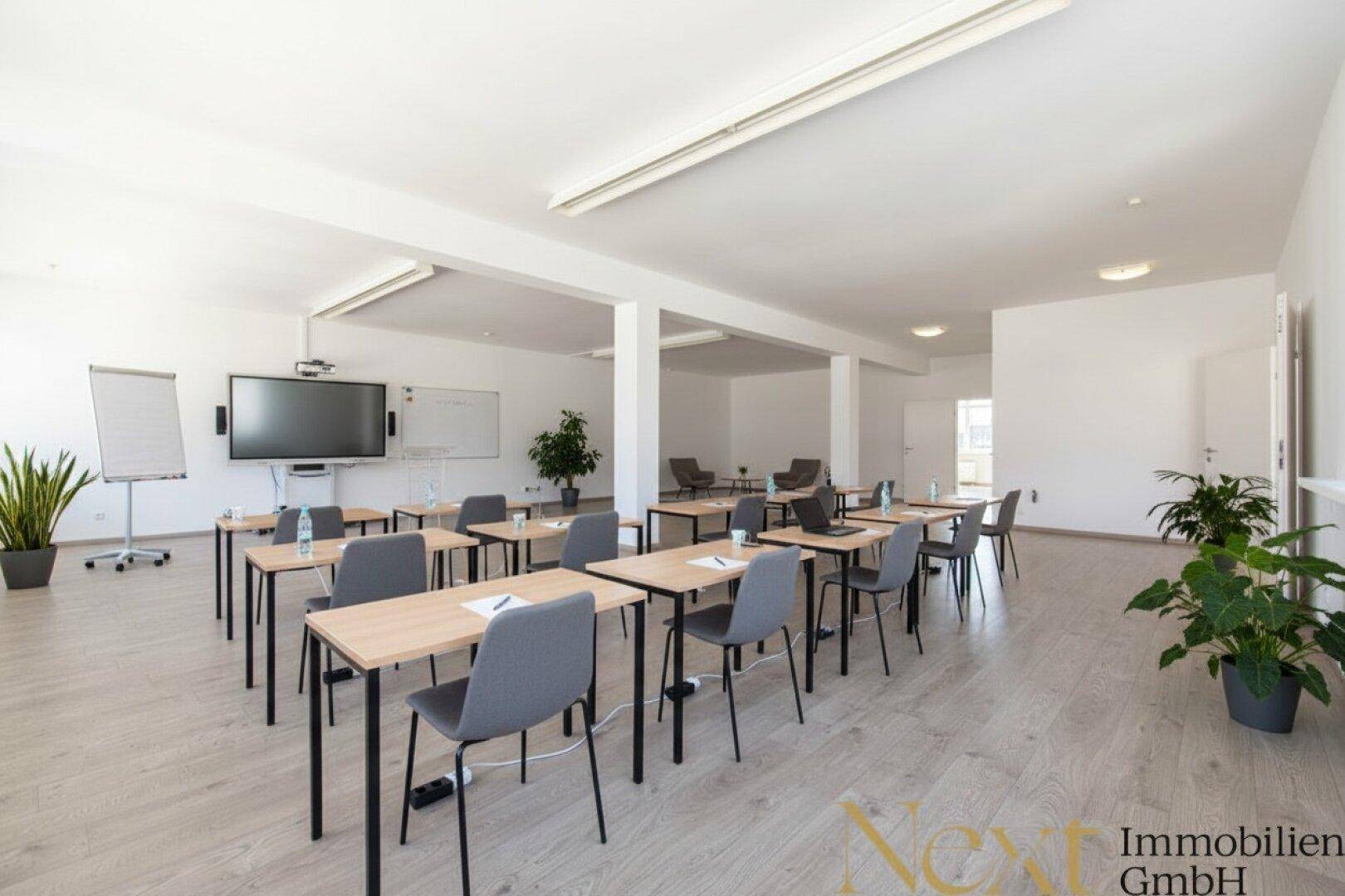 Moderner Konferenzraum mit flexibler Bestuhlung, großem Bildschirm und Pflanzen, ideal für Schulungen und Meetings.