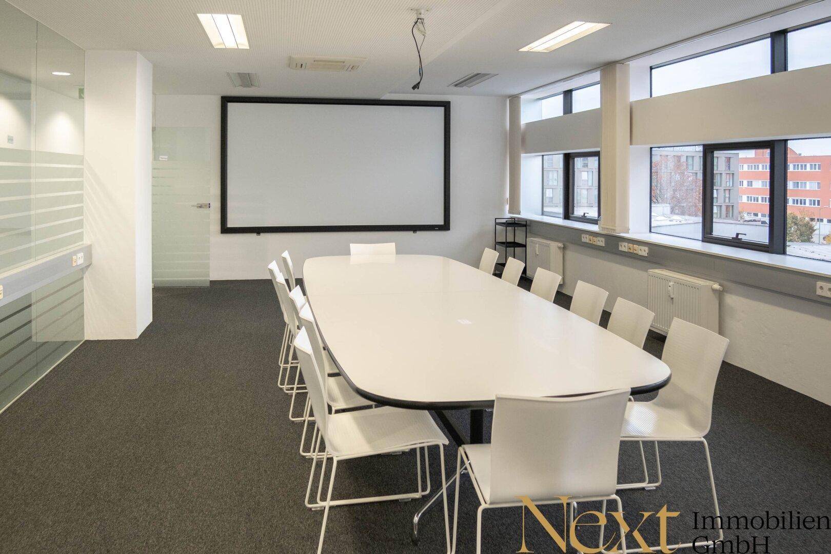 Geräumiger Konferenzraum mit großem weißen Tisch, bequemen Stühlen und einem Projektionsbildschirm für Präsentationen.
