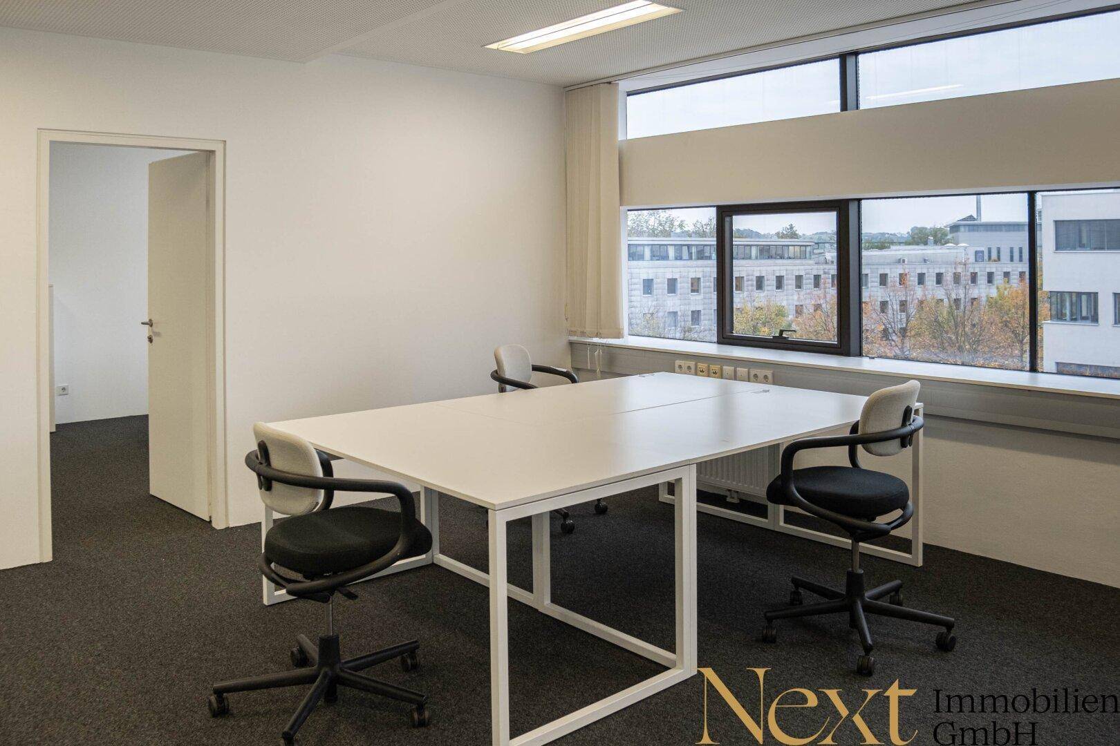 Einzelner Bürobereich mit modernem weißen Schreibtisch, ergonomischen Stühlen und Blick nach draußen durch große Fenster.