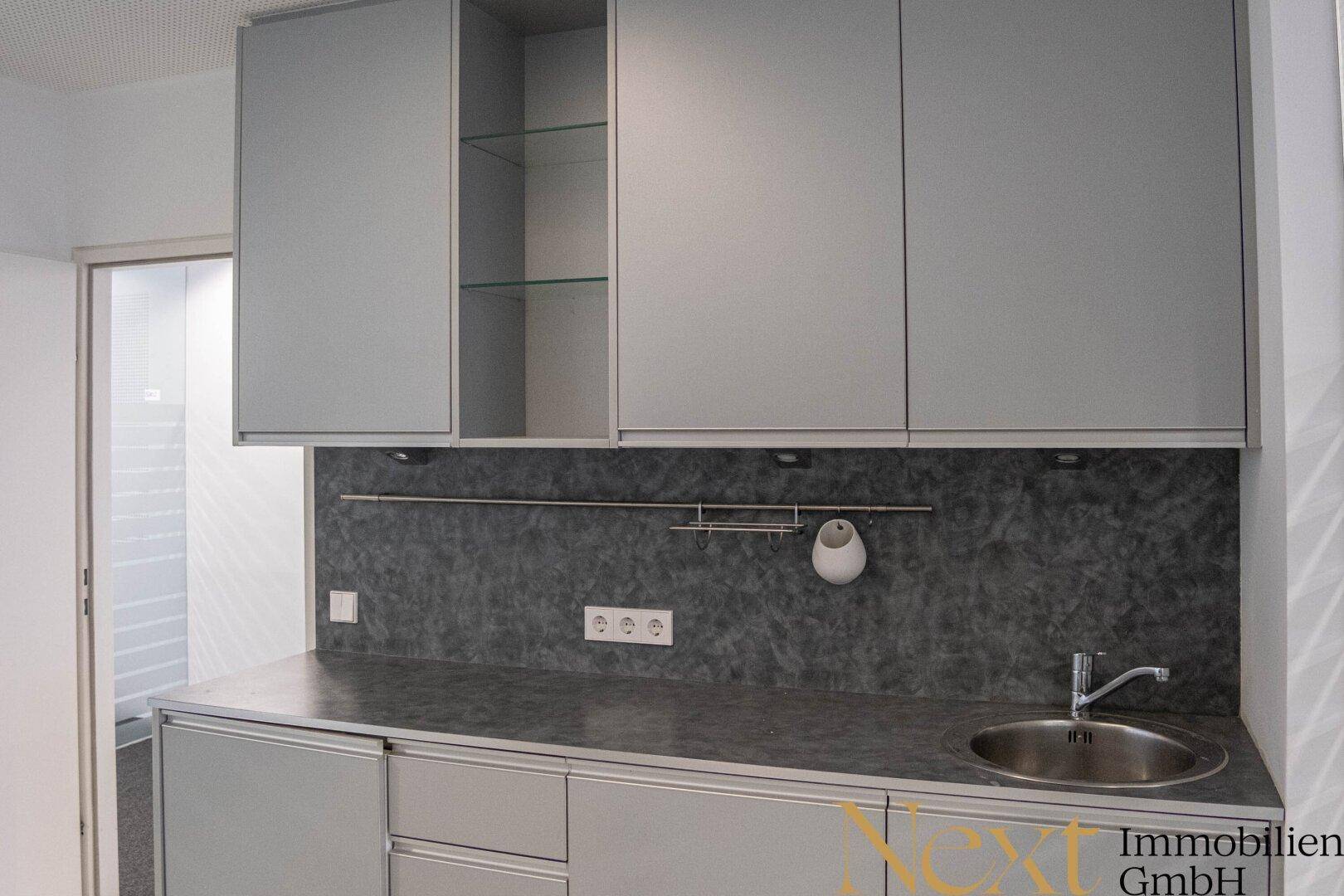 Funktionale Küchenzeile mit grauen Schränken, Spüle und dunkler Arbeitsplatte, ideal für kleine Büros oder Teeküchen.