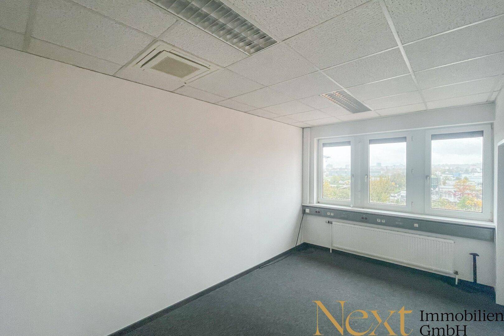 Leerer Büroraum mit Teppichboden, großen Fenstern und Blick auf die Umgebung.