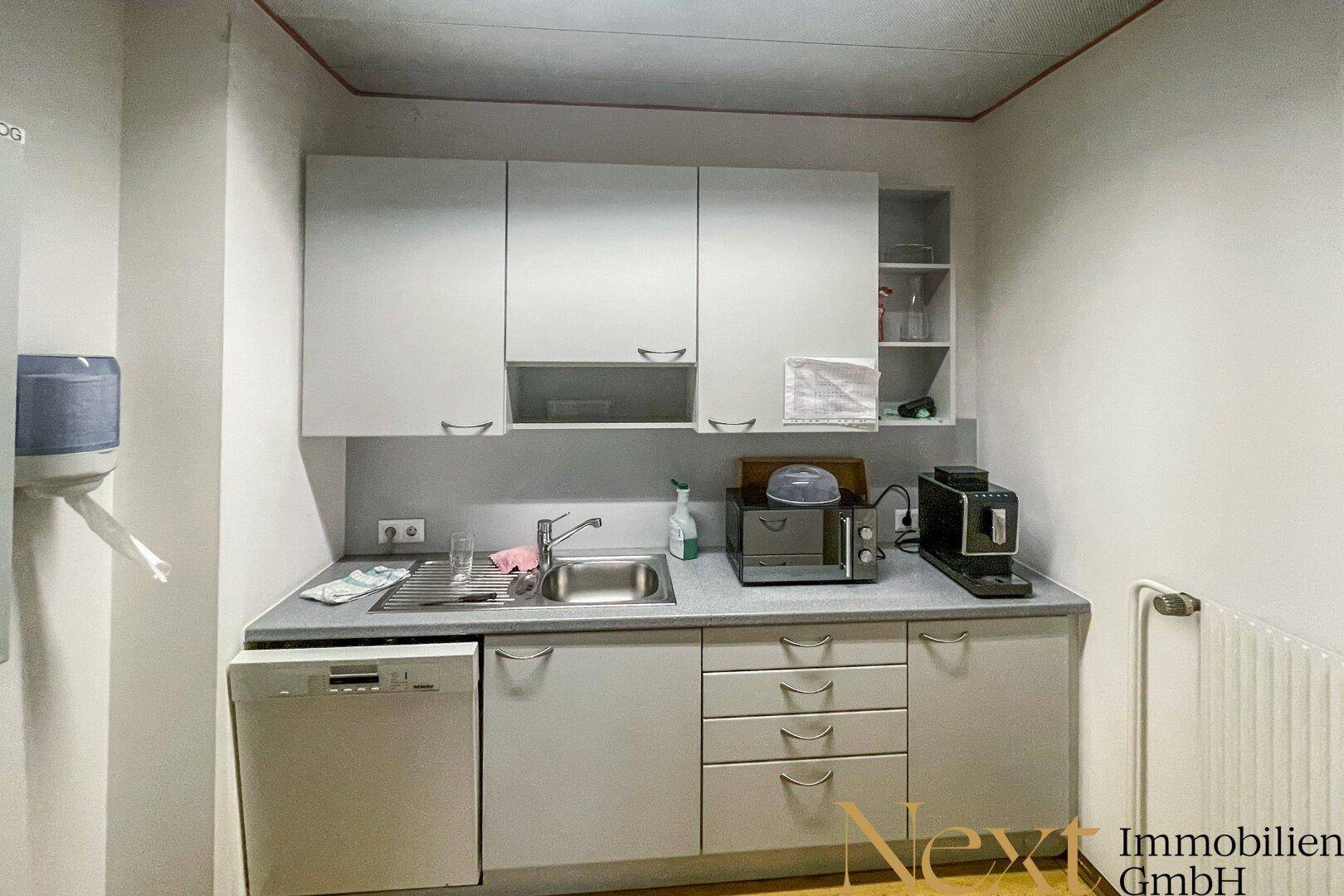 Kleine Küchenzeile mit weißen Schränken, Spüle, Geschirrspüler und Kaffeemaschine.