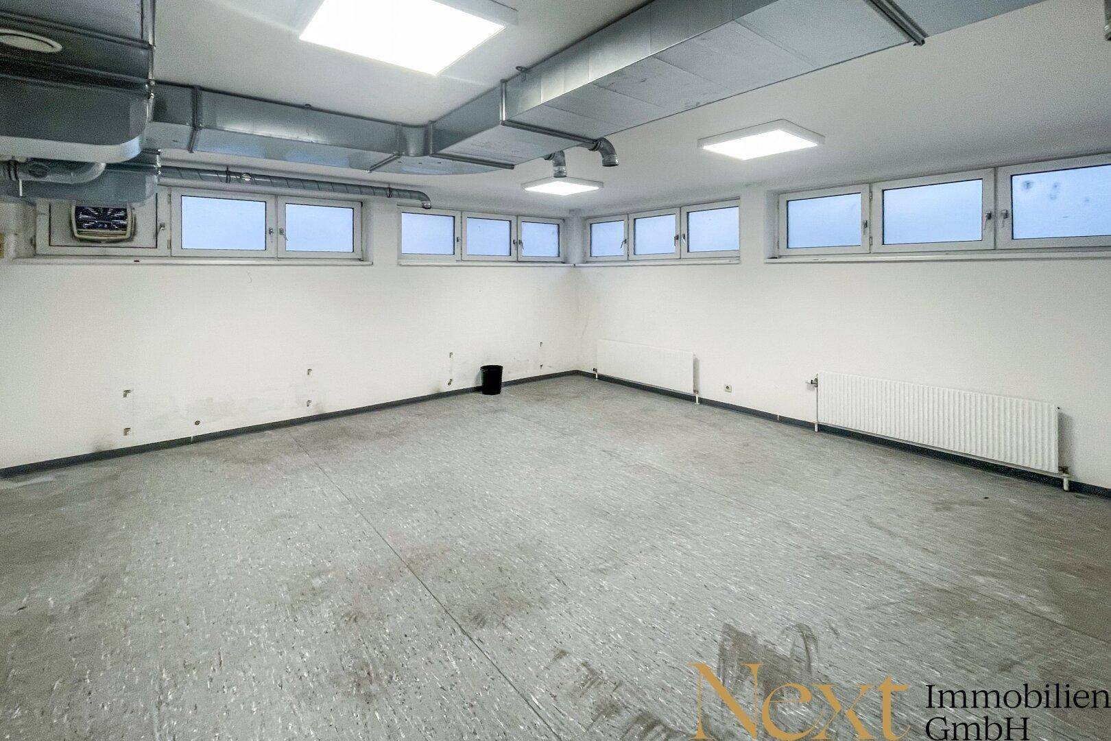 Leerer Raum mit vielen kleinen Fenstern, Heizkörpern und sichtbaren Lüftungsrohren an der Decke.