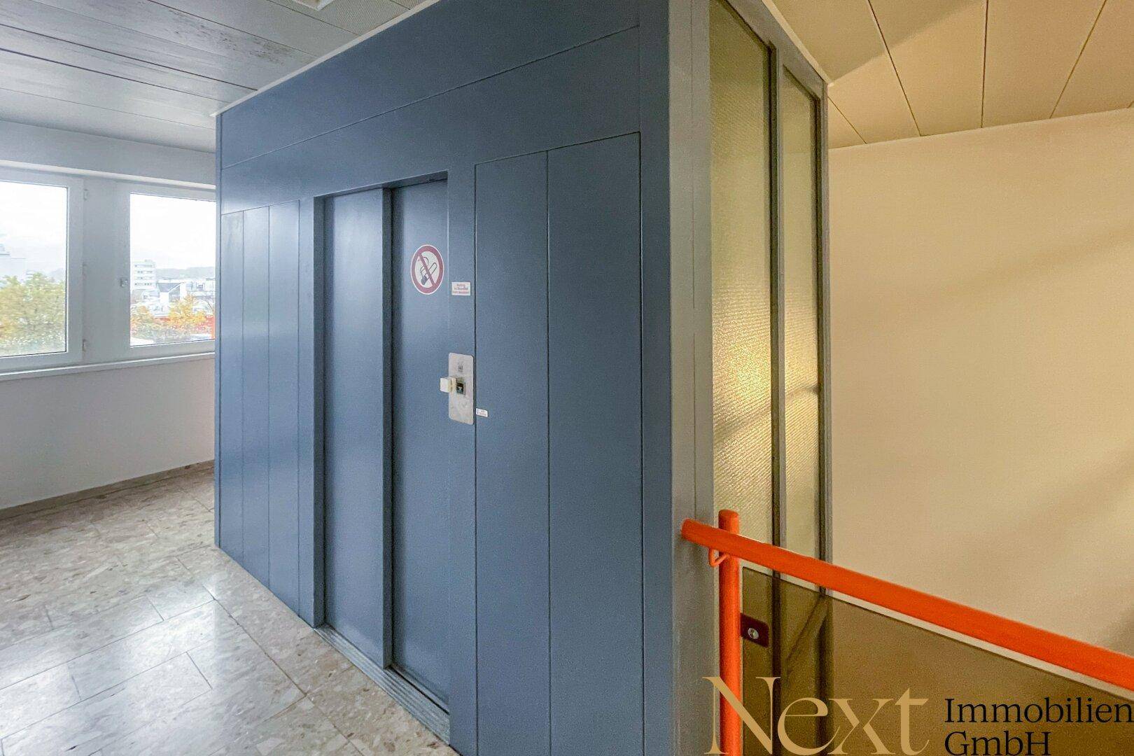 Flur mit einem modernen blauen Aufzug und Treppenhaus mit orangefarbenem Geländer.