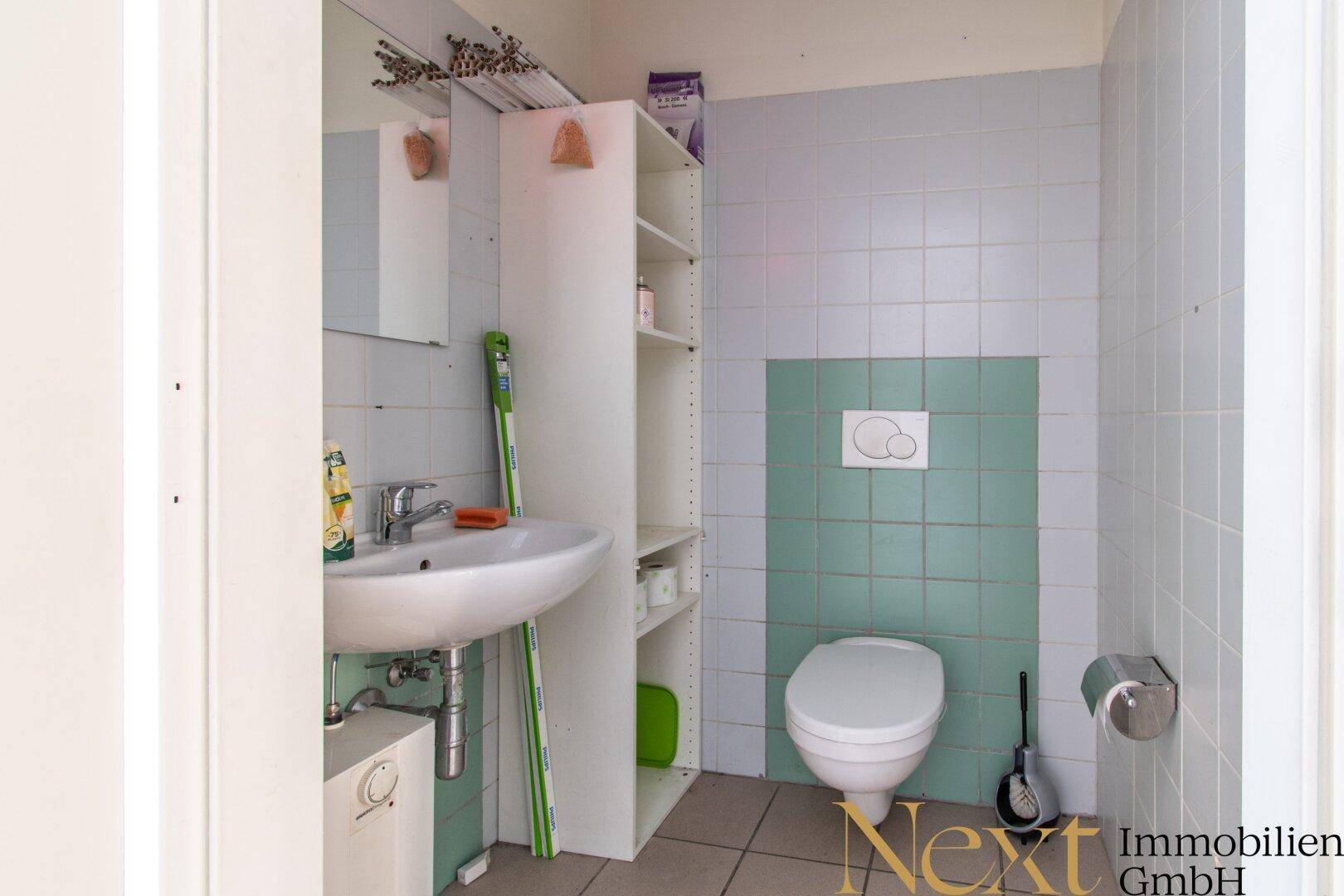 Einfaches Badezimmer mit WC, Waschbecken und hellblau-grünen Fliesen, ausgestattet mit einem Regal.