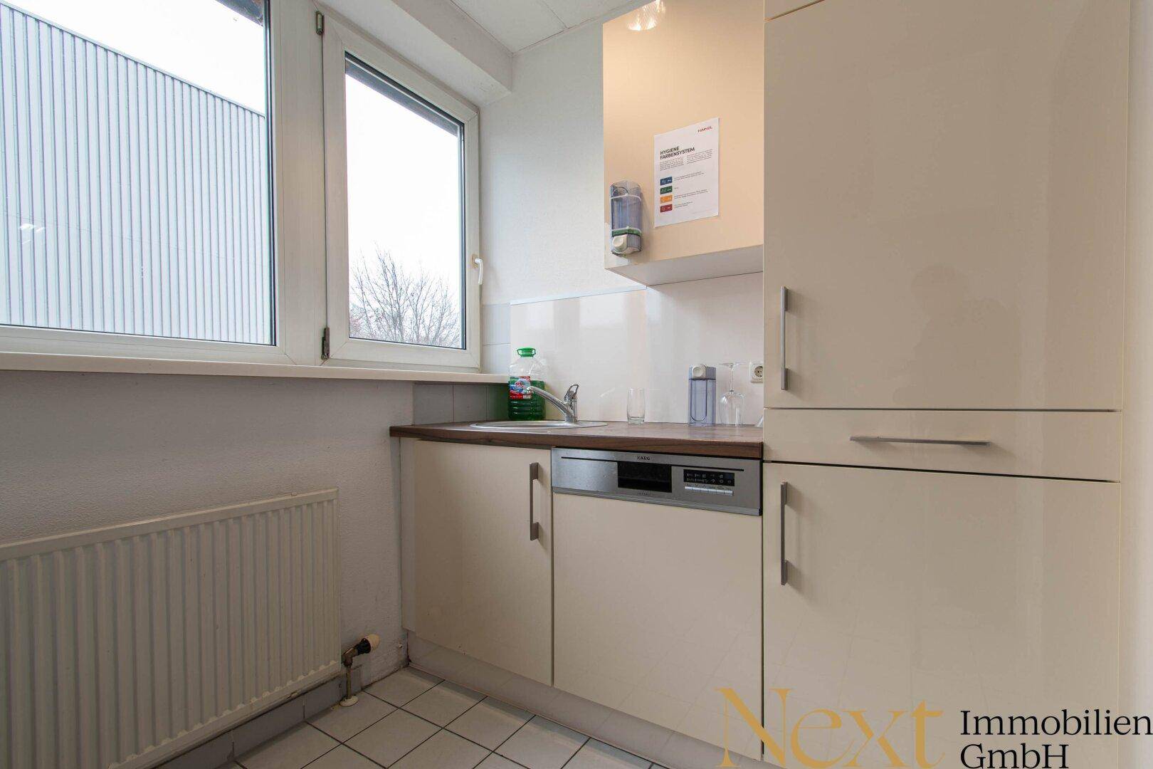 Kleine Küche mit beigen Schränken, Spülmaschine und Fenster, ideal für eine Teeküche oder Pausenraum.