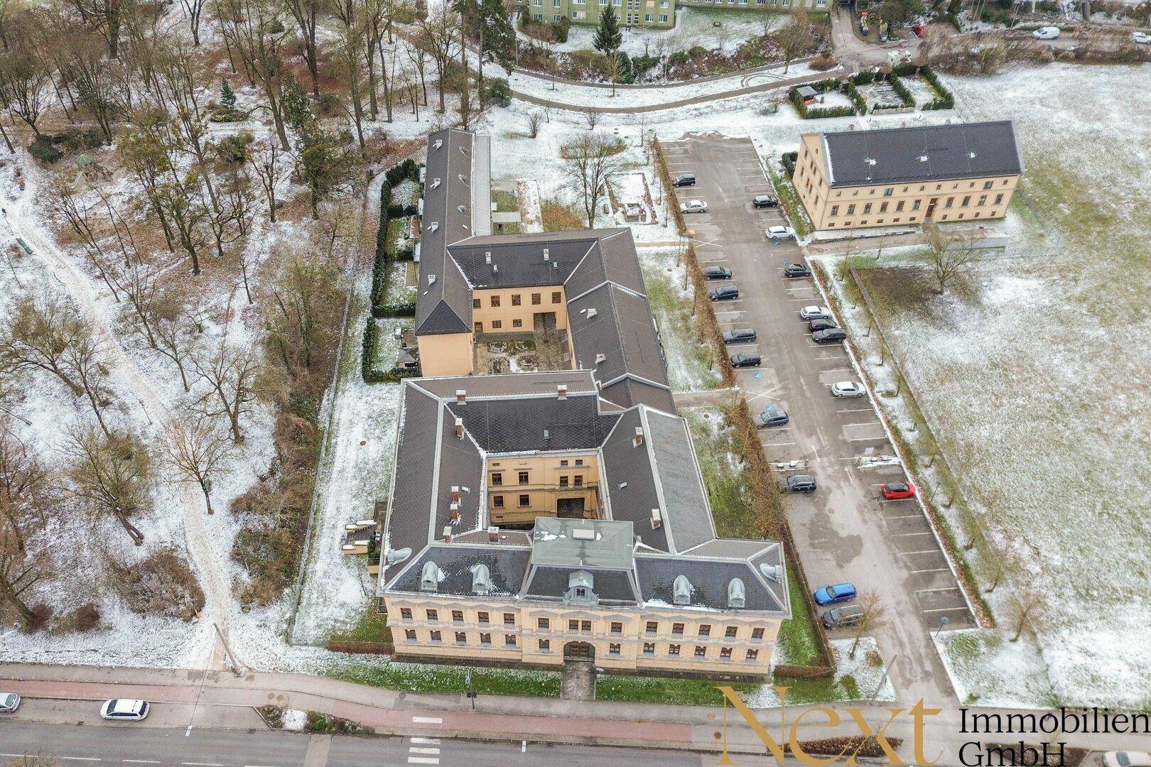 Weitläufige Luftaufnahme des historischen Gebäudes und seiner Umgebung mit Parkplätzen und schneebedeckten Flächen.