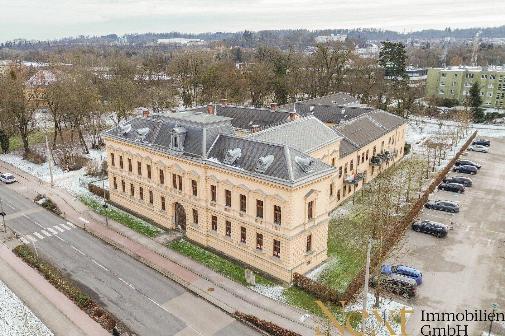 Luftaufnahme des weitläufigen historischen Gebäudekomplexes mit umliegenden Parkplätzen und Grünflächen im Winter.