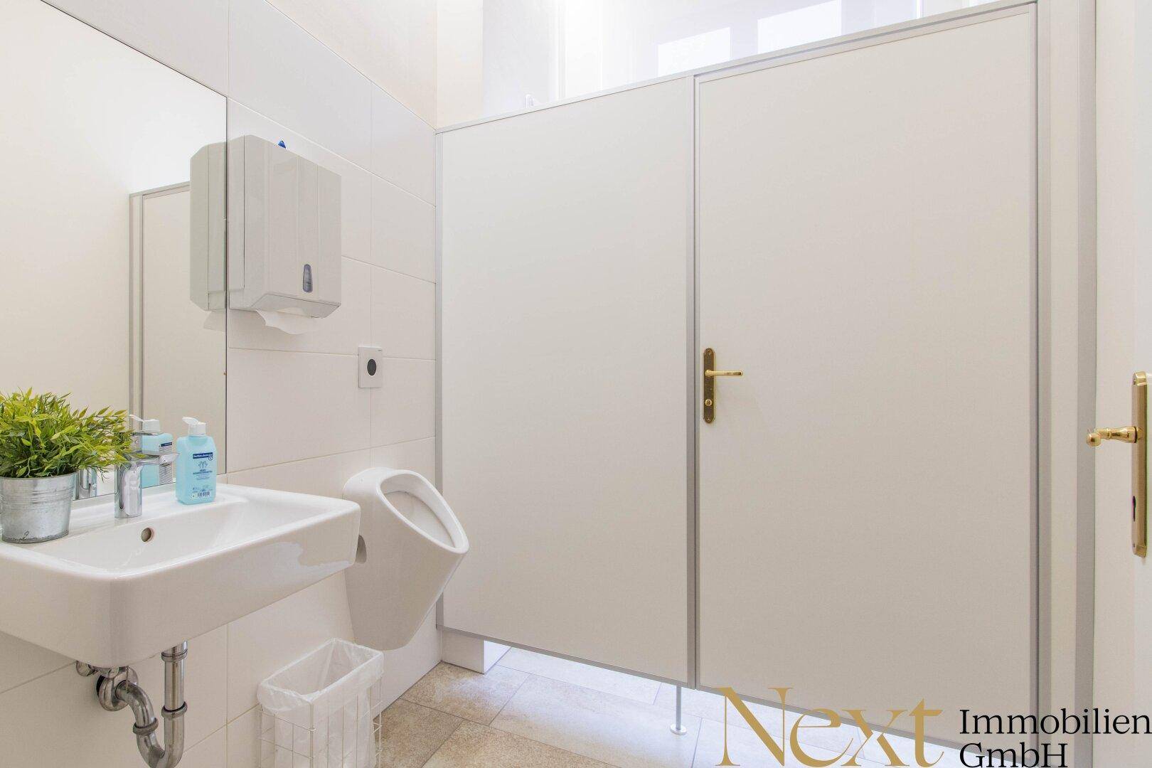 Saubere und funktionale öffentliche Toilette mit Urinal, Waschbecken und weißen Trennwänden.