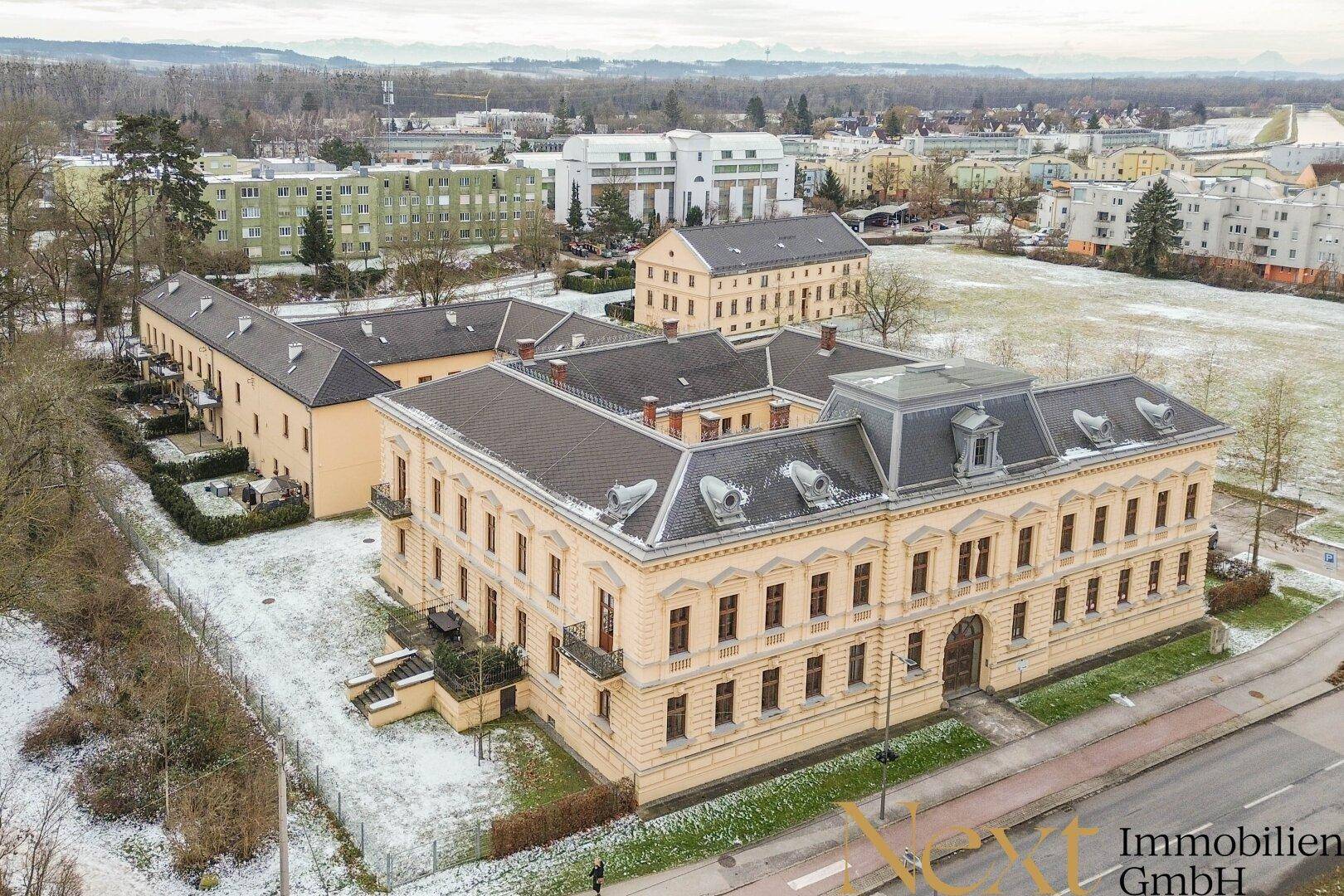 Luftaufnahme eines historischen Gebäudekomplexes mit umliegenden Grünflächen und leichtem Schneefall.