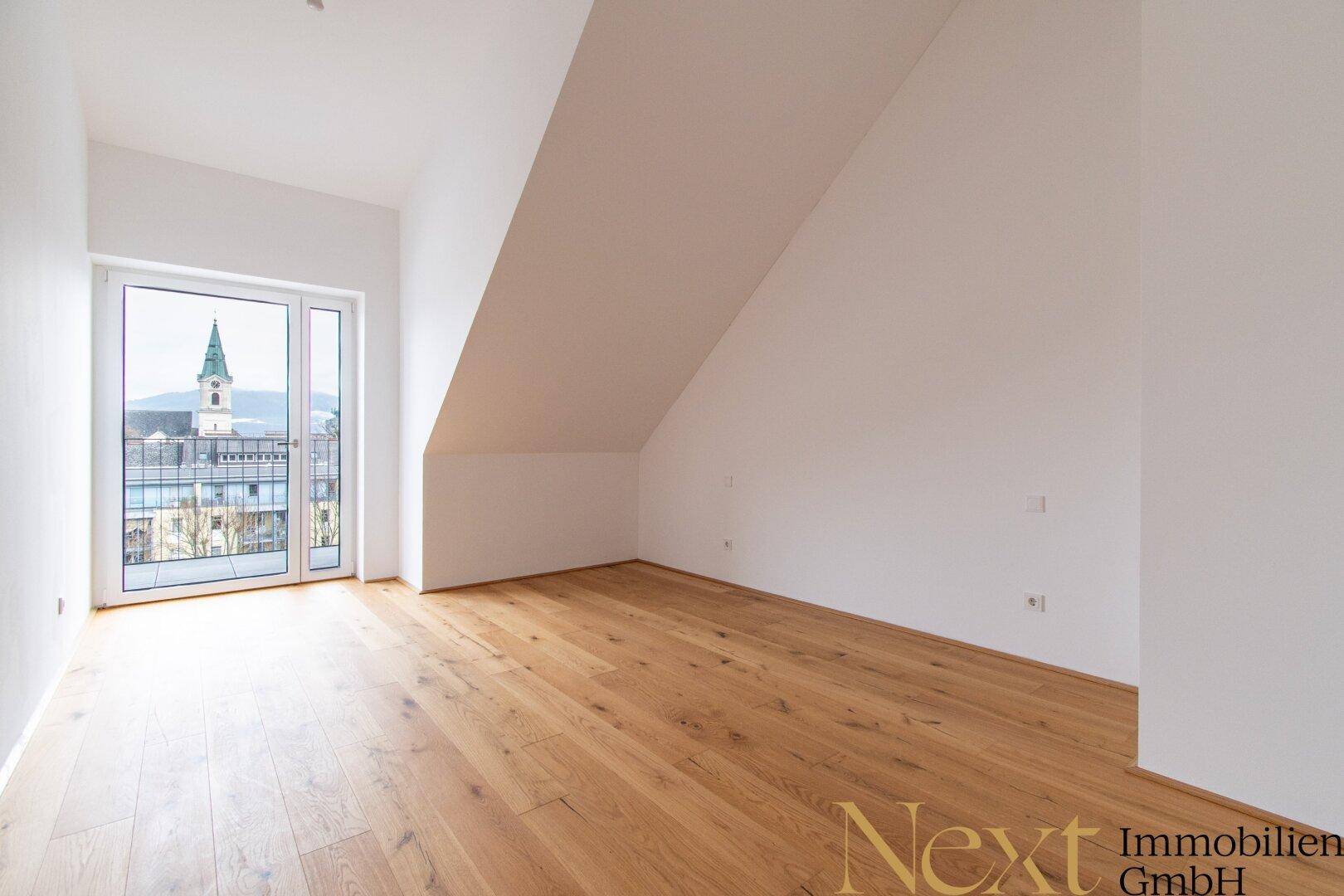 Leeres Schlafzimmer mit Holzboden, Dachschräge und Zugang zu einem Balkon.