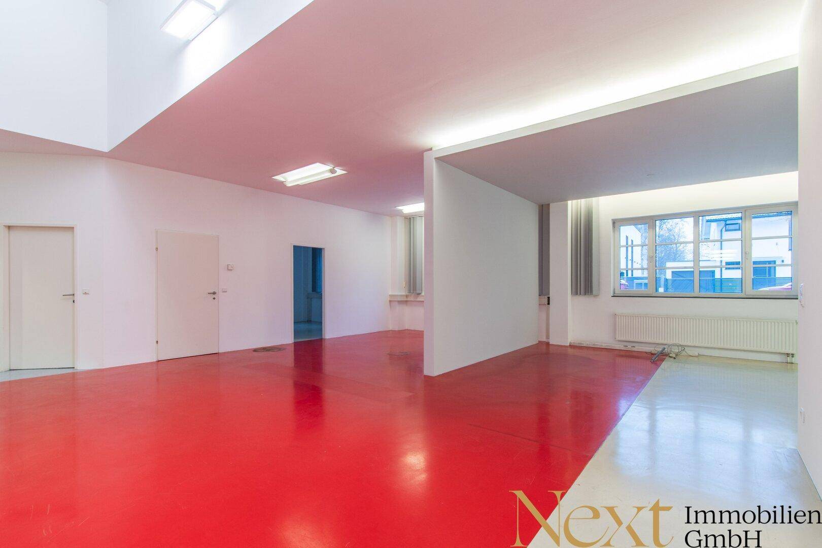 Großer, offener Bürobereich mit auffälligem roten Boden, Fenstern und mehreren Türen.