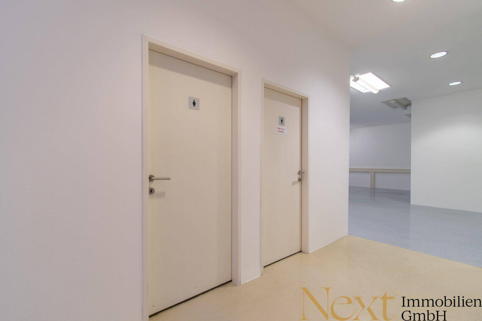 Heller Flur mit zwei Türen zu den Toiletten und Blick in einen weiteren Raum.